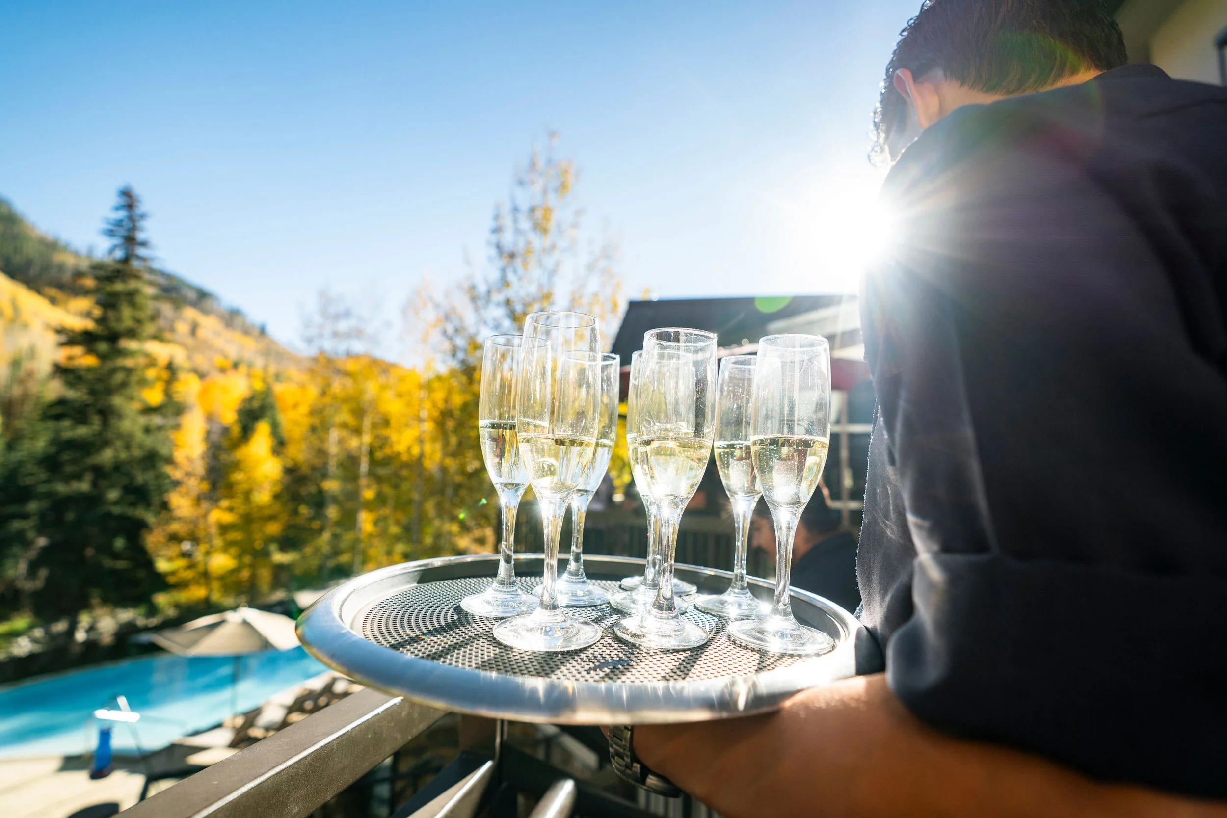 Person holding a tray with six glasses of champagne outdoors on a sunny day, with trees and a mountain in the background.