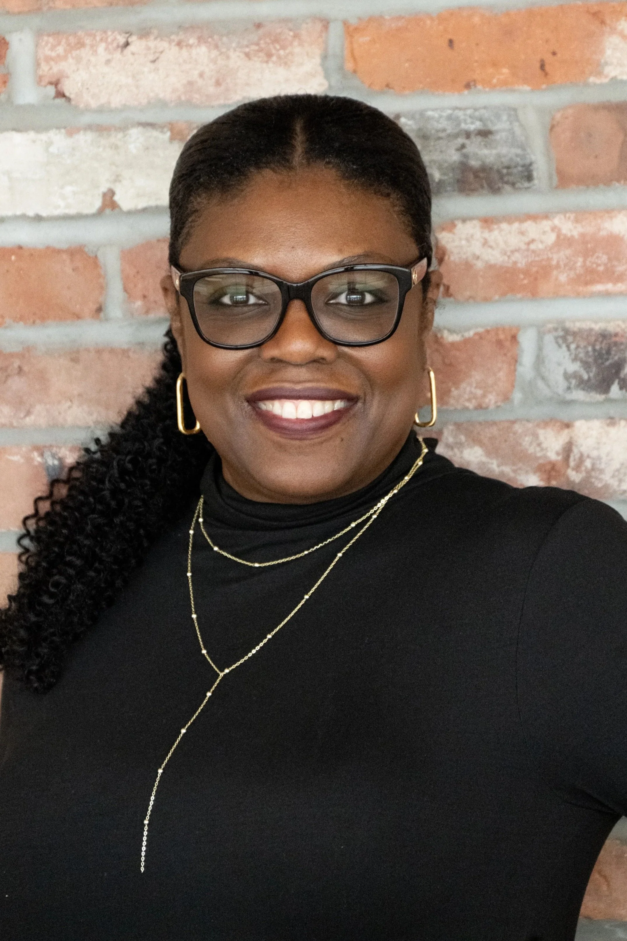 A smiling woman with glasses and gold jewelry stands against a brick wall.