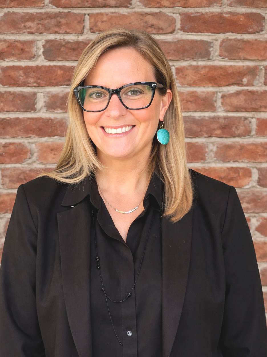 A smiling woman with shoulder-length blonde hair wearing black glasses, a black blazer, black shirt, turquoise earrings, a silver necklace, and a black beaded necklace stands in front of a brick wall.