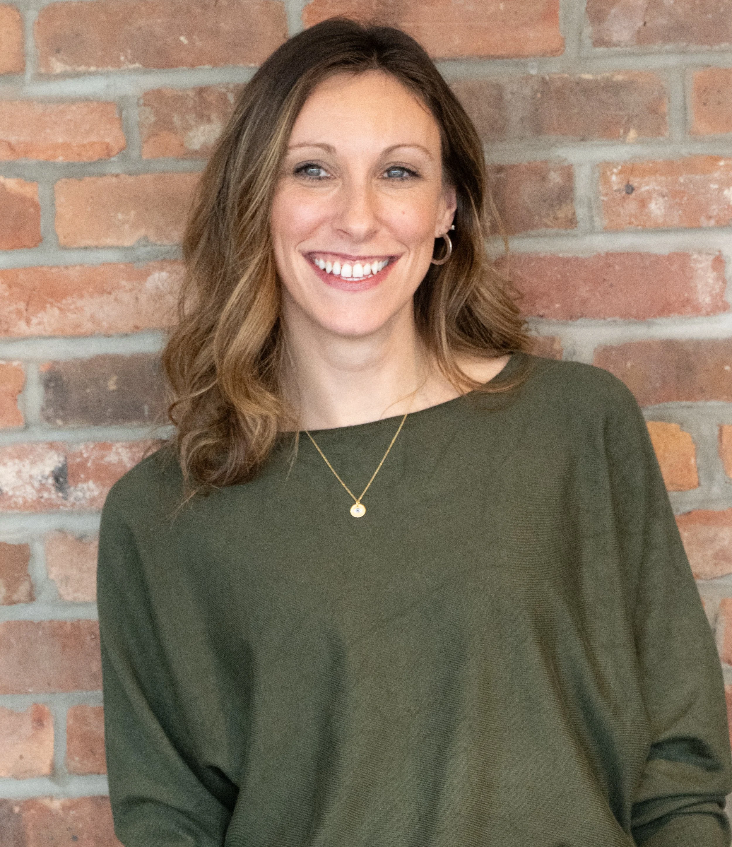 A woman with shoulder-length wavy brown hair, smiling, standing in front of a brick wall, wearing a dark green top and a small necklace.