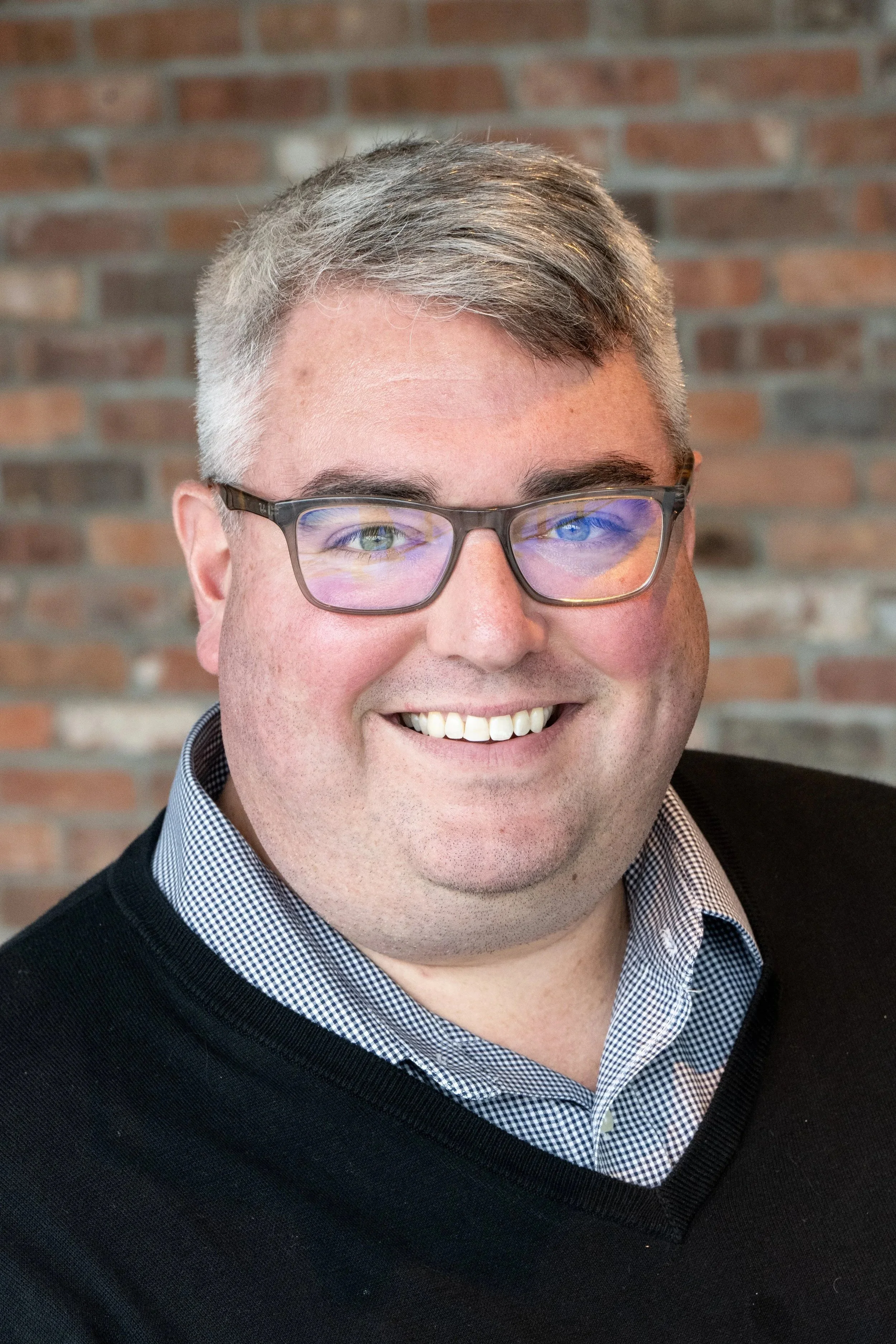 A smiling man with glasses and short gray hair, wearing a black sweater over a blue and white checkered shirt, standing in front of a brick wall.