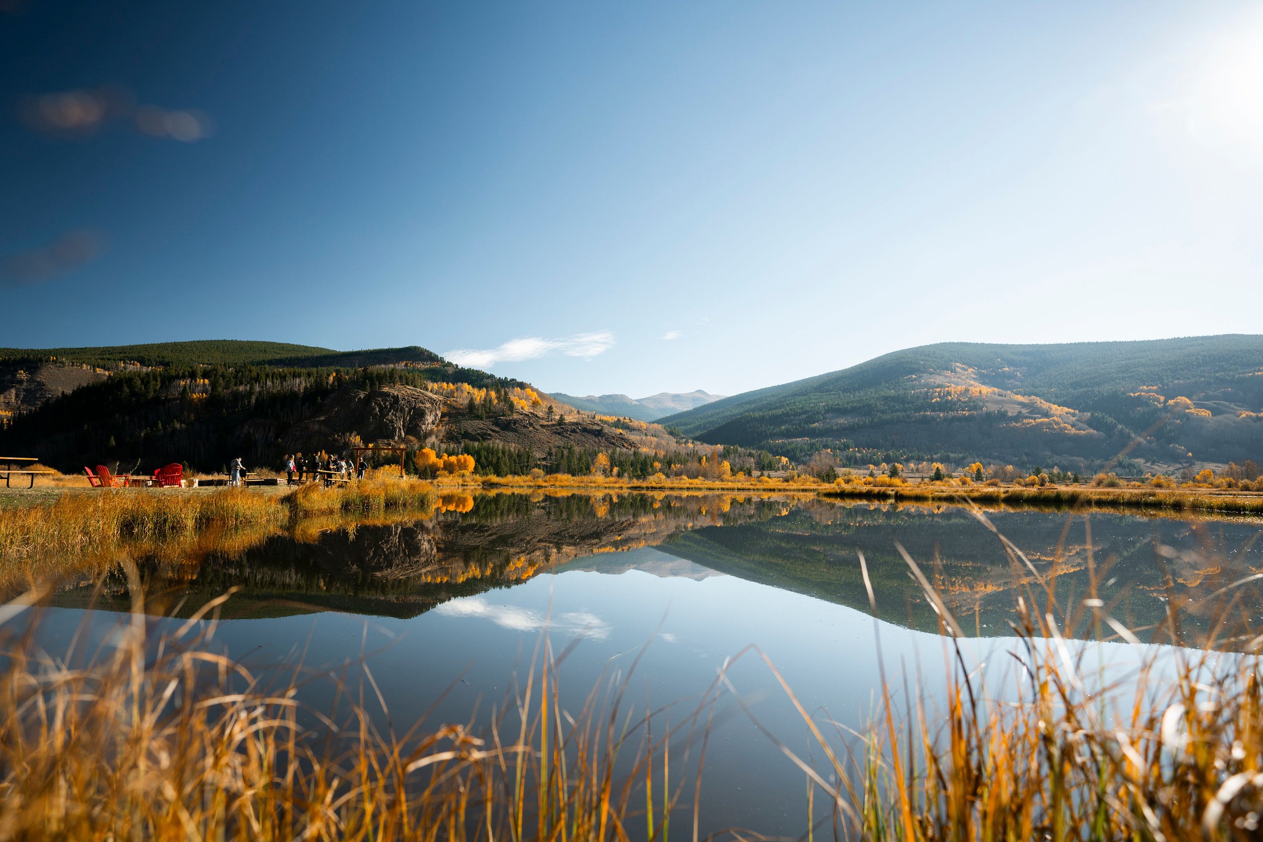 A serene mountain landscape with a calm lake reflecting the blue sky, green hills, and autumn-colored trees.