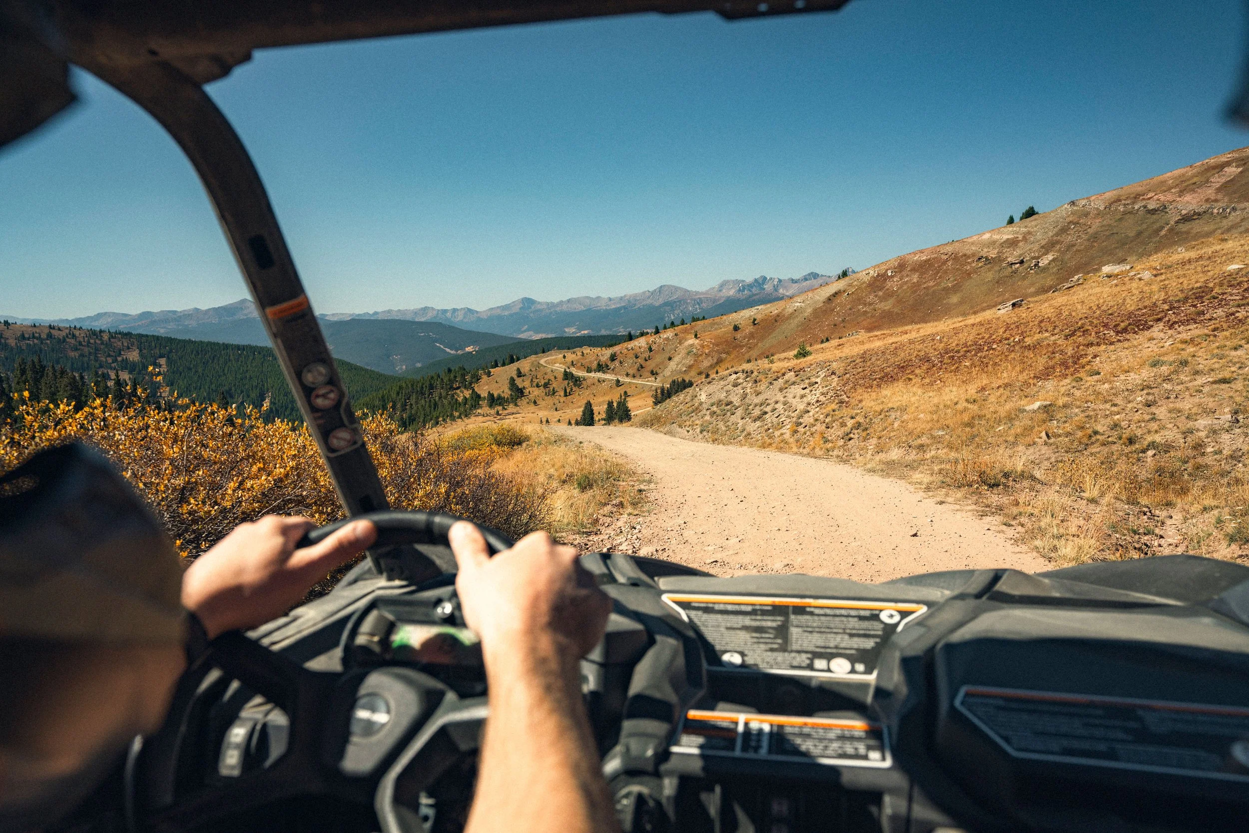A person driving an off-road vehicle on a dirt trail through a hilly, mountainous landscape with sparse trees and dry grass under a clear blue sky.