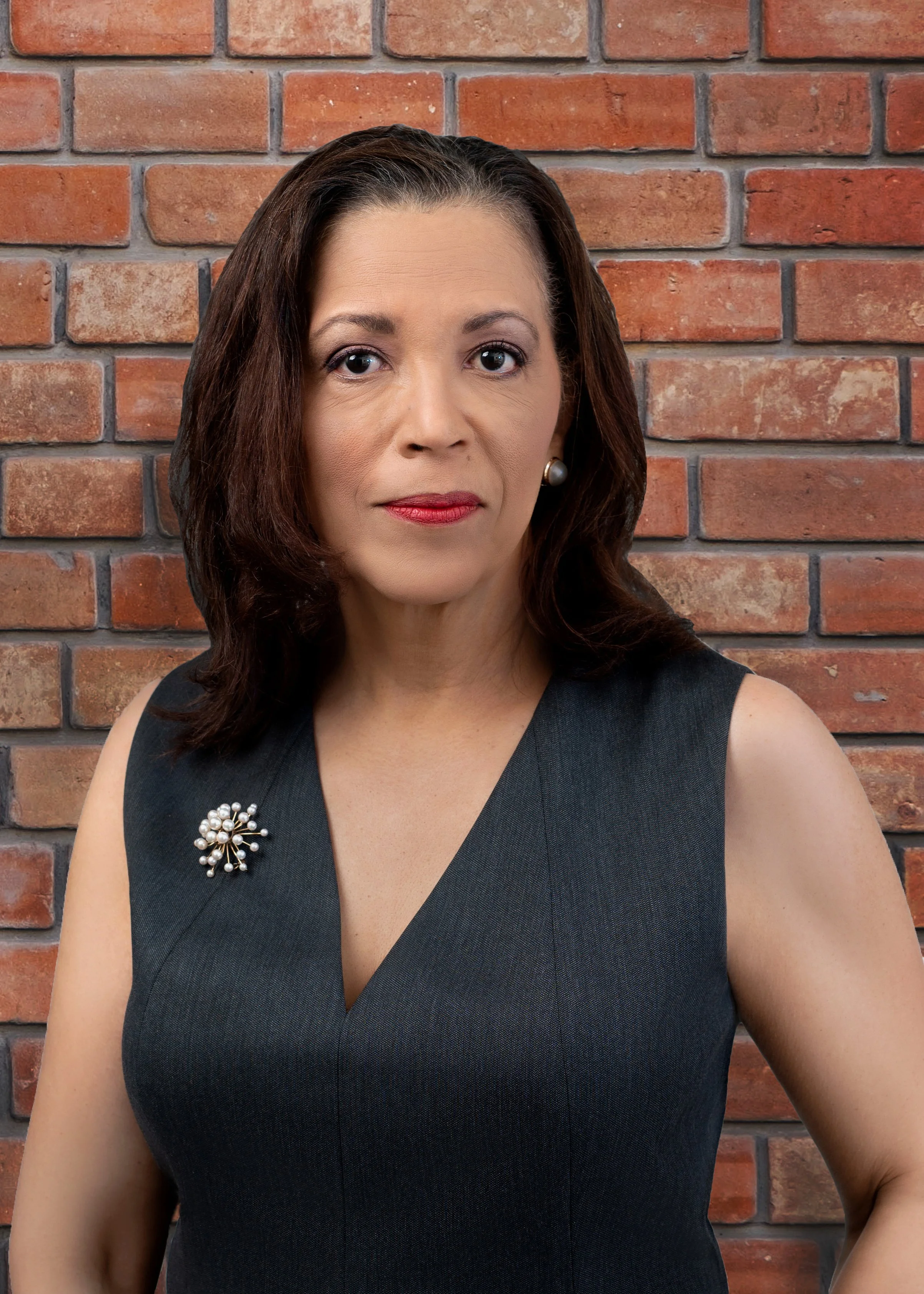A woman with dark hair wearing a black sleeveless top with a pearl brooch on the shoulder, standing in front of a brick wall.