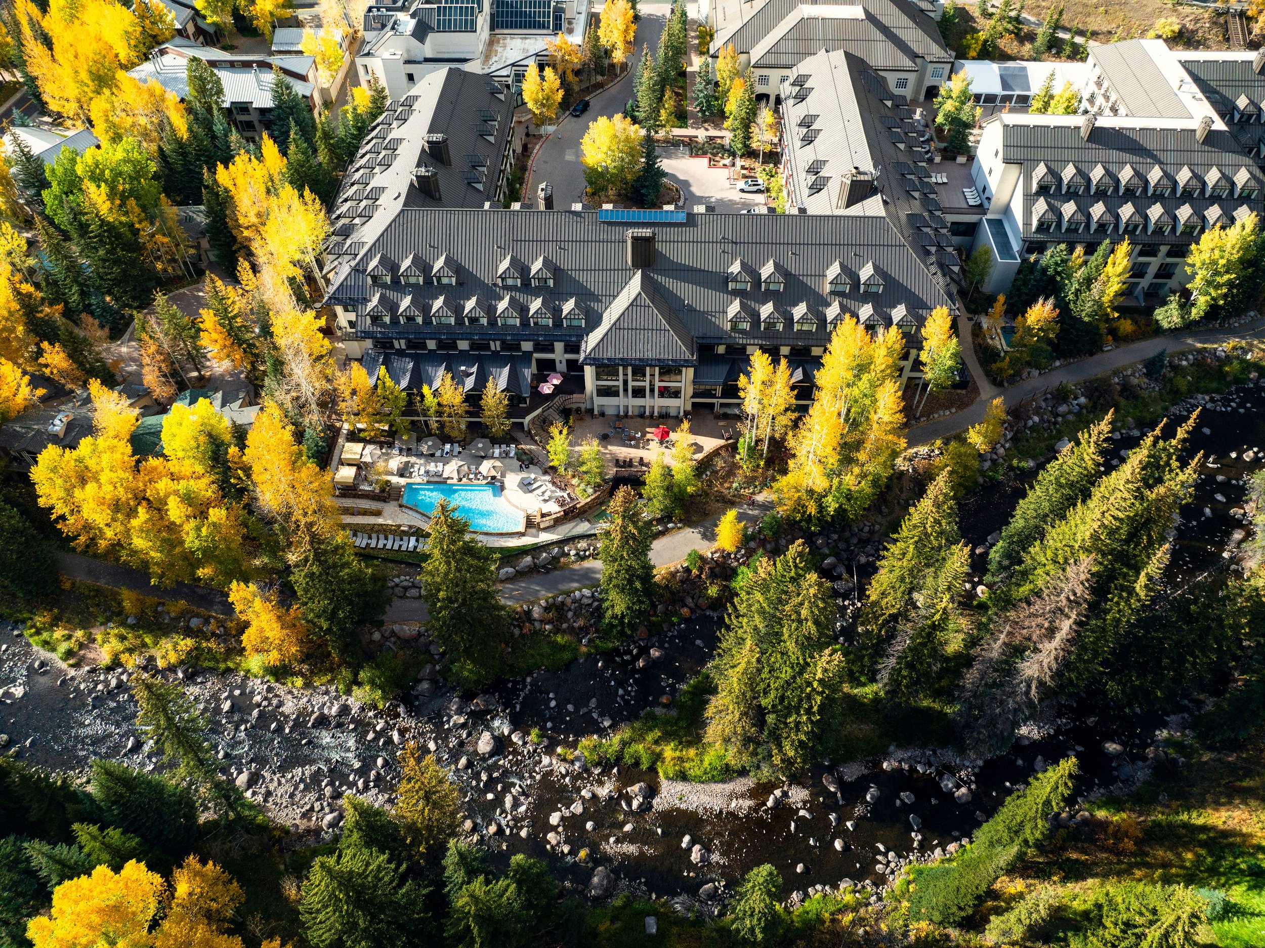 Aerial view of a residential area with houses, trees showcasing fall colors, a swimming pool, and a river running through a wooded landscape.