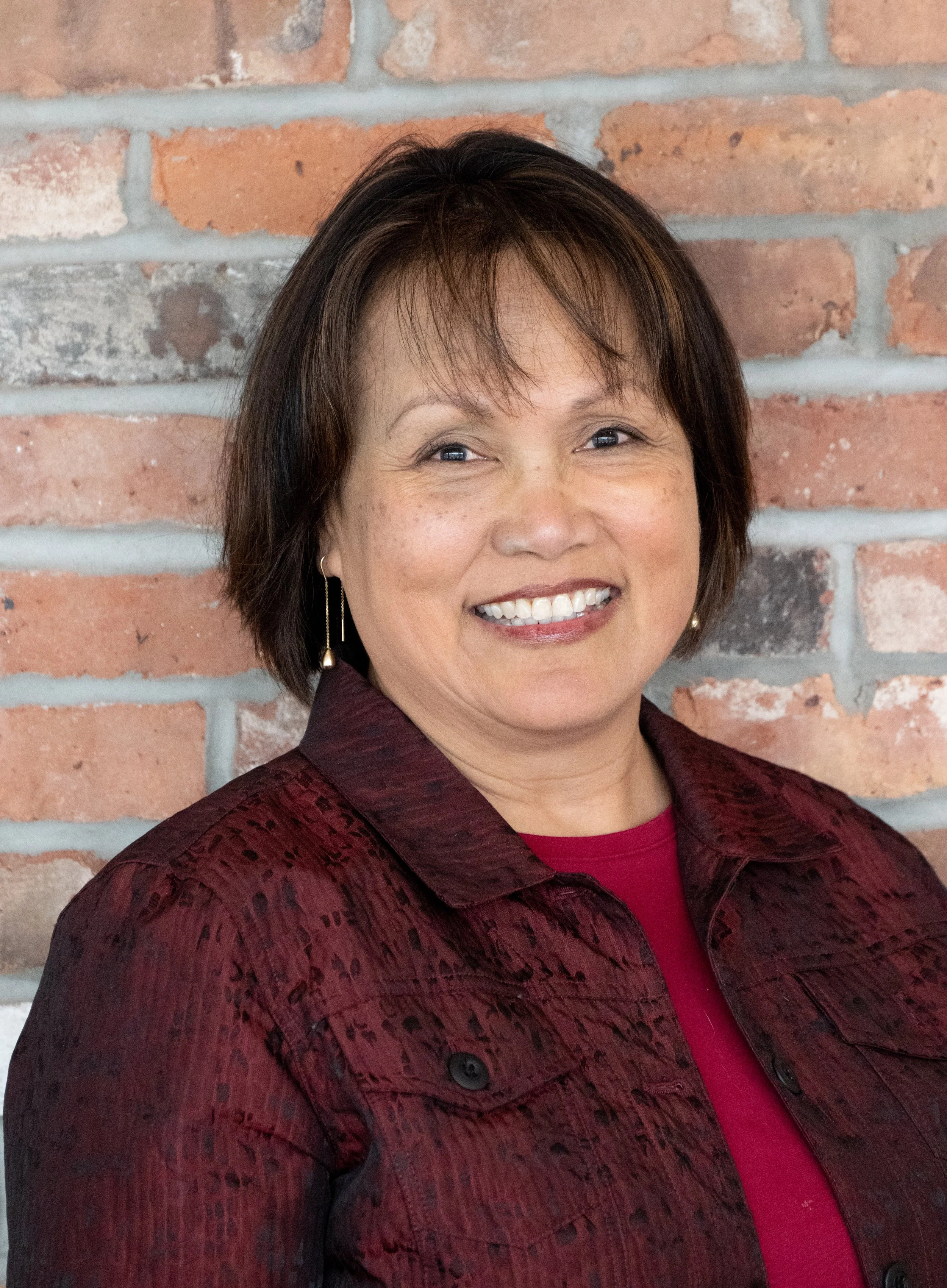 A woman with short dark brown hair wearing a dark red patterned jacket over a red top, smiling in front of a brick wall.