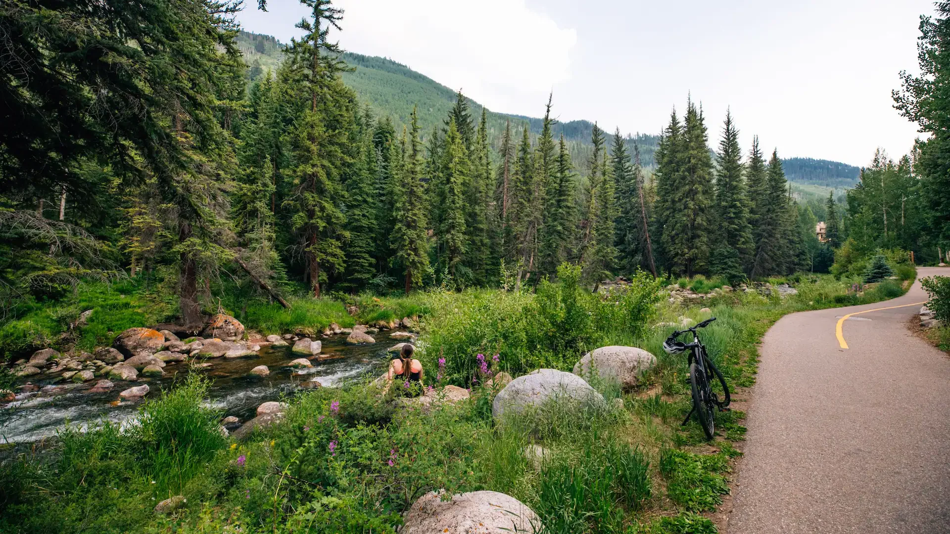 A person sits beside a river in a lush forested mountain landscape, with a bicycle parked on a winding road nearby.