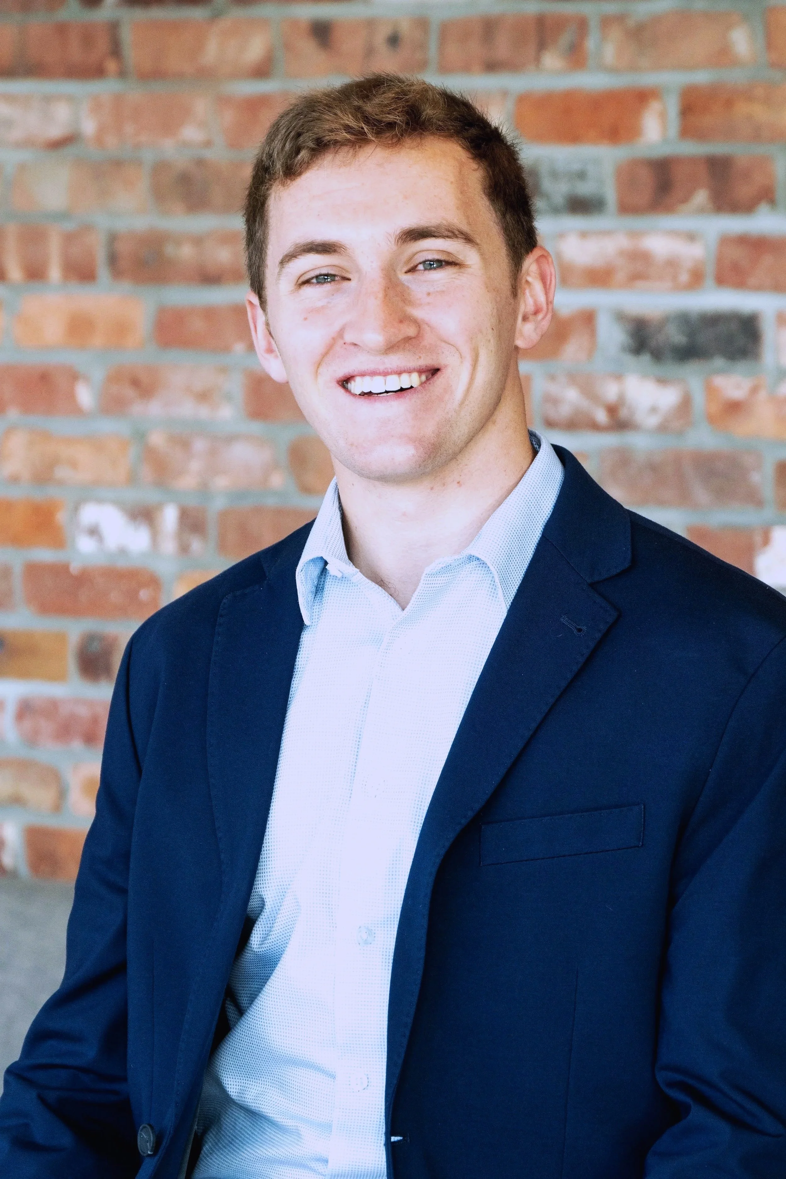 A young man with short brown hair, blue eyes, and a cheerful smile, wearing a navy blue blazer and light blue dress shirt, standing in front of a brick wall.