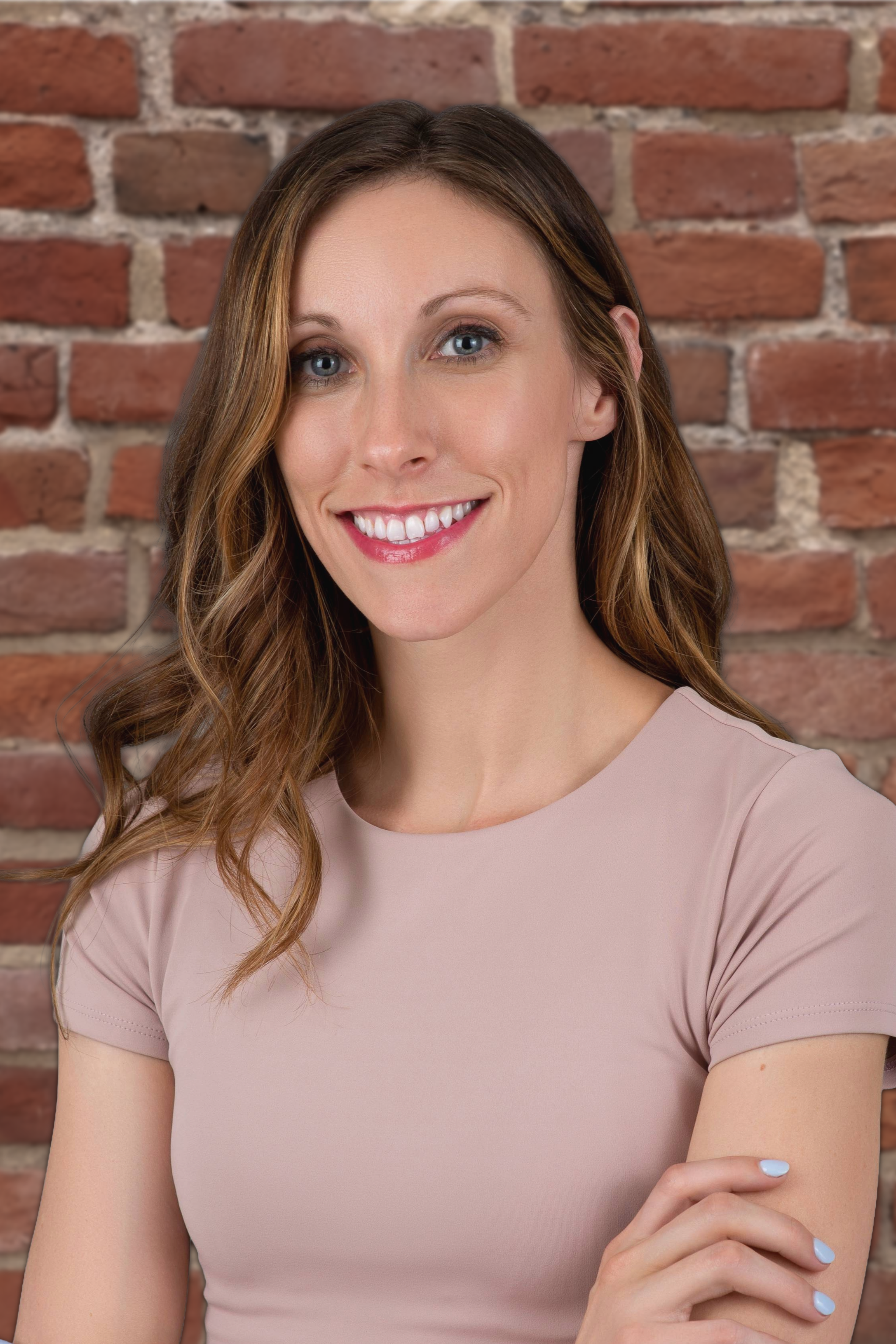A woman with shoulder-length wavy brown hair, smiling, standing in front of a brick wall, wearing a dark green top and a small necklace.