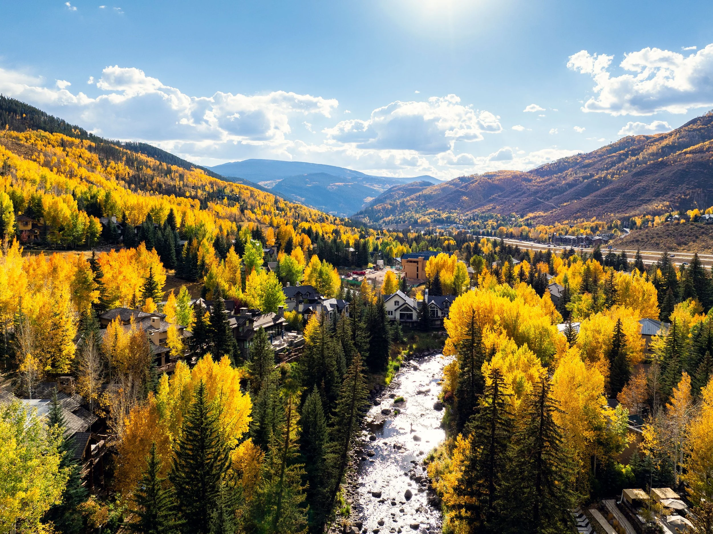 A scenic mountain valley with a river running through a forest of colorful autumn trees, houses, and distant mountains under a partly cloudy sky.