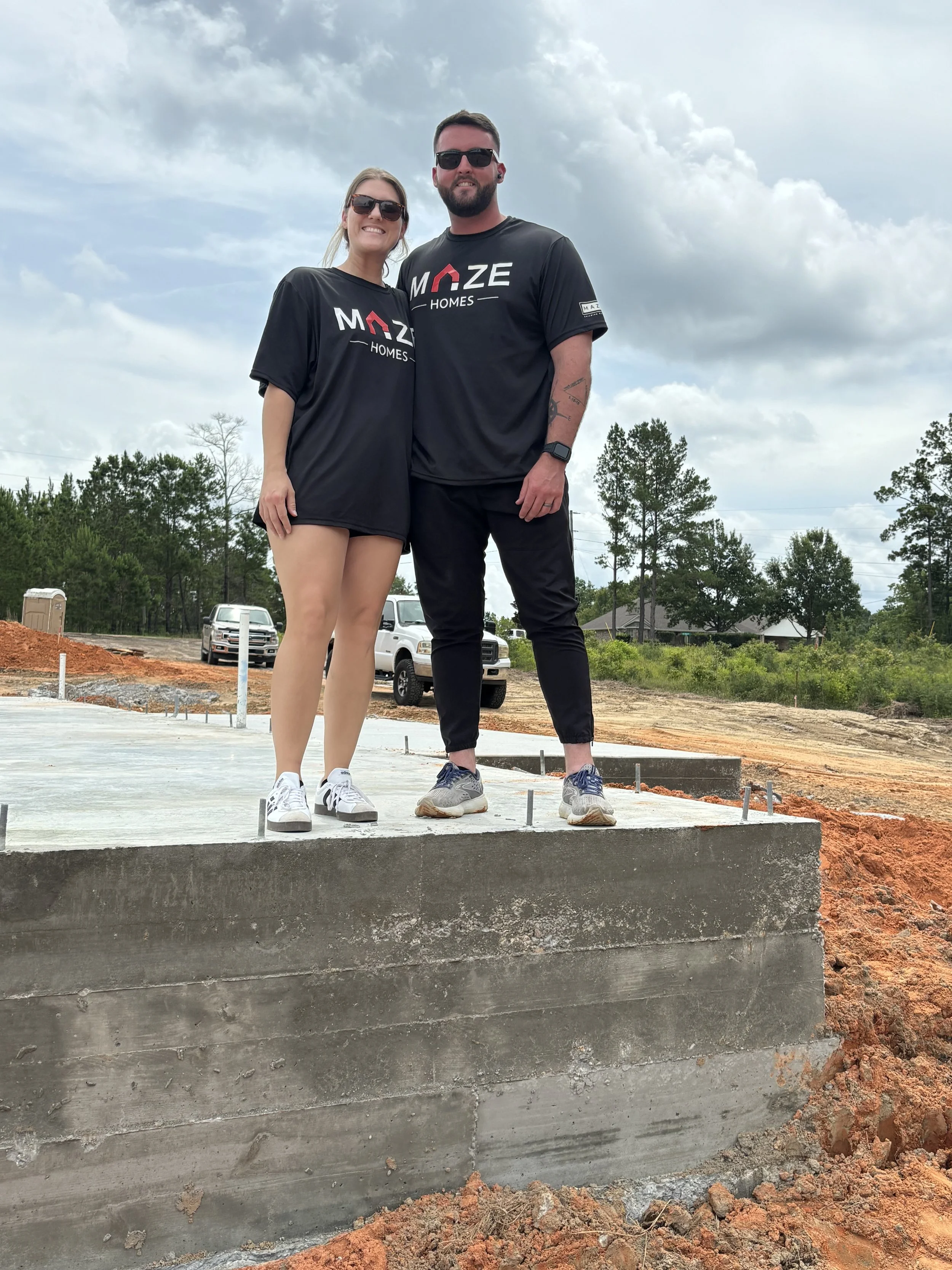 A smiling man and woman wearing matching black t-shirts with 'MAZE HOMES' logo, standing on a concrete foundation at a construction site with trees and vehicles in the background.