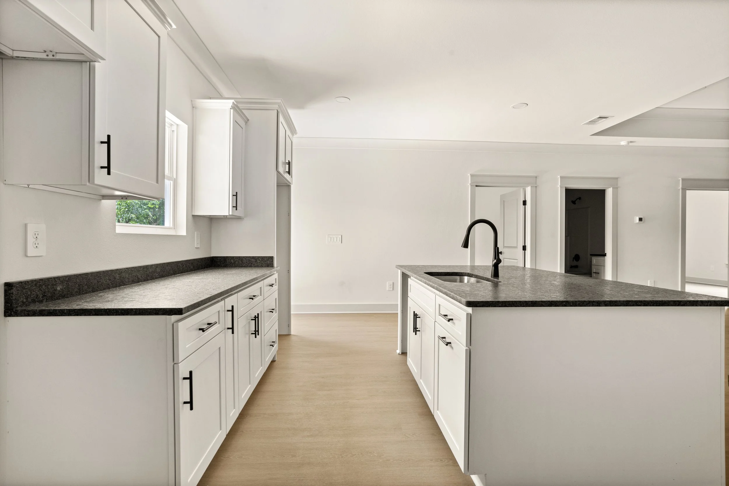 Empty kitchen with white cabinets, black handles, black countertops, and a kitchen island with a black faucet, featuring natural light from a window.