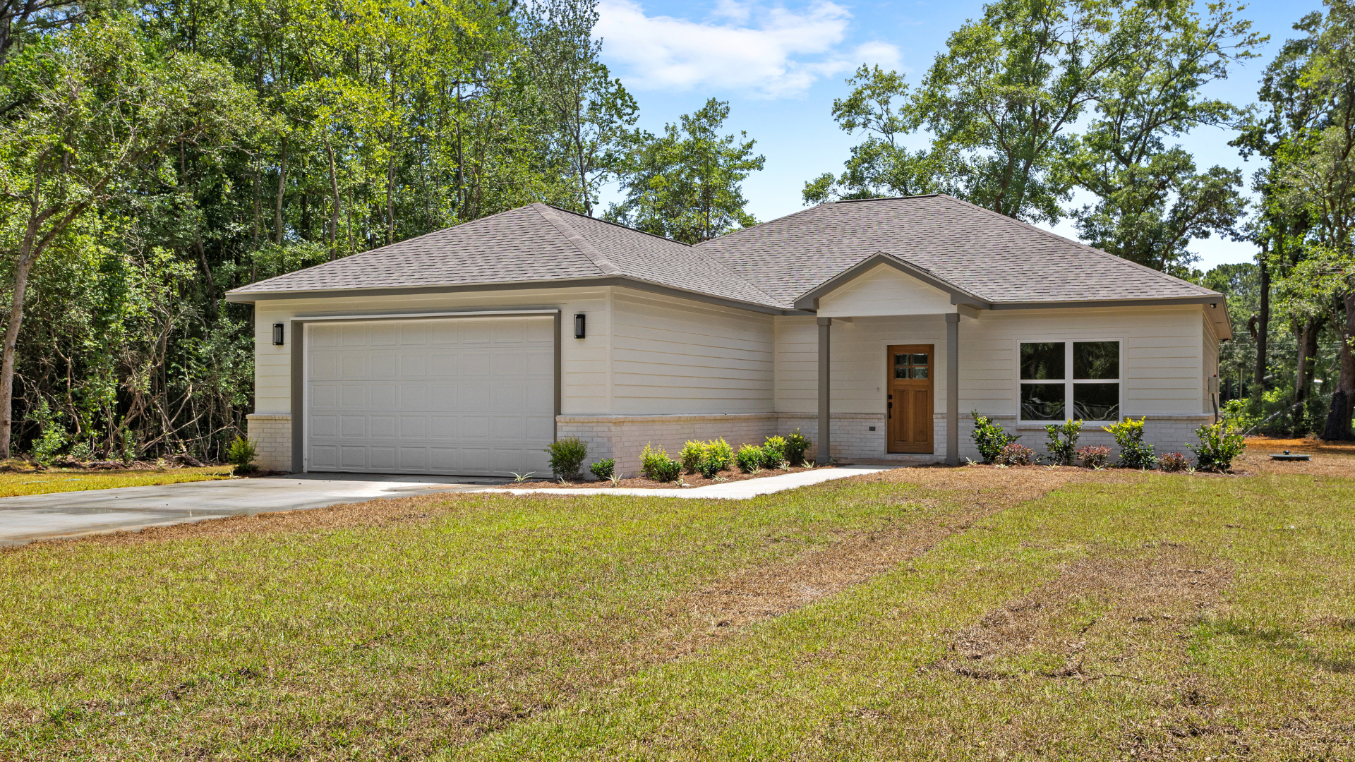 New single-story house with white siding, brown front door, and attached garage, surrounded by a lawn and trees in the background.