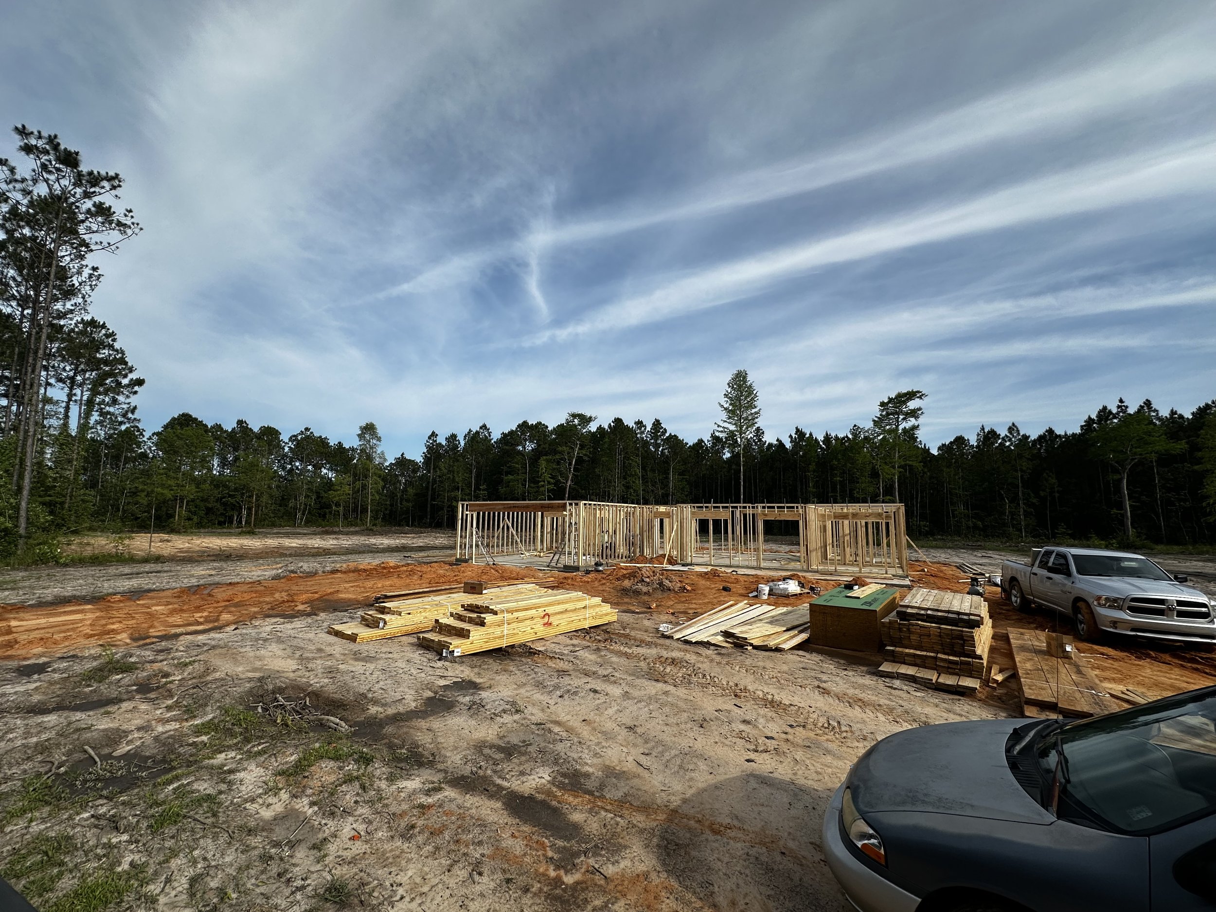 Construction site with wooden framing, stacks of lumber, a pickup truck, and a car, with a forest and blue sky in the background.