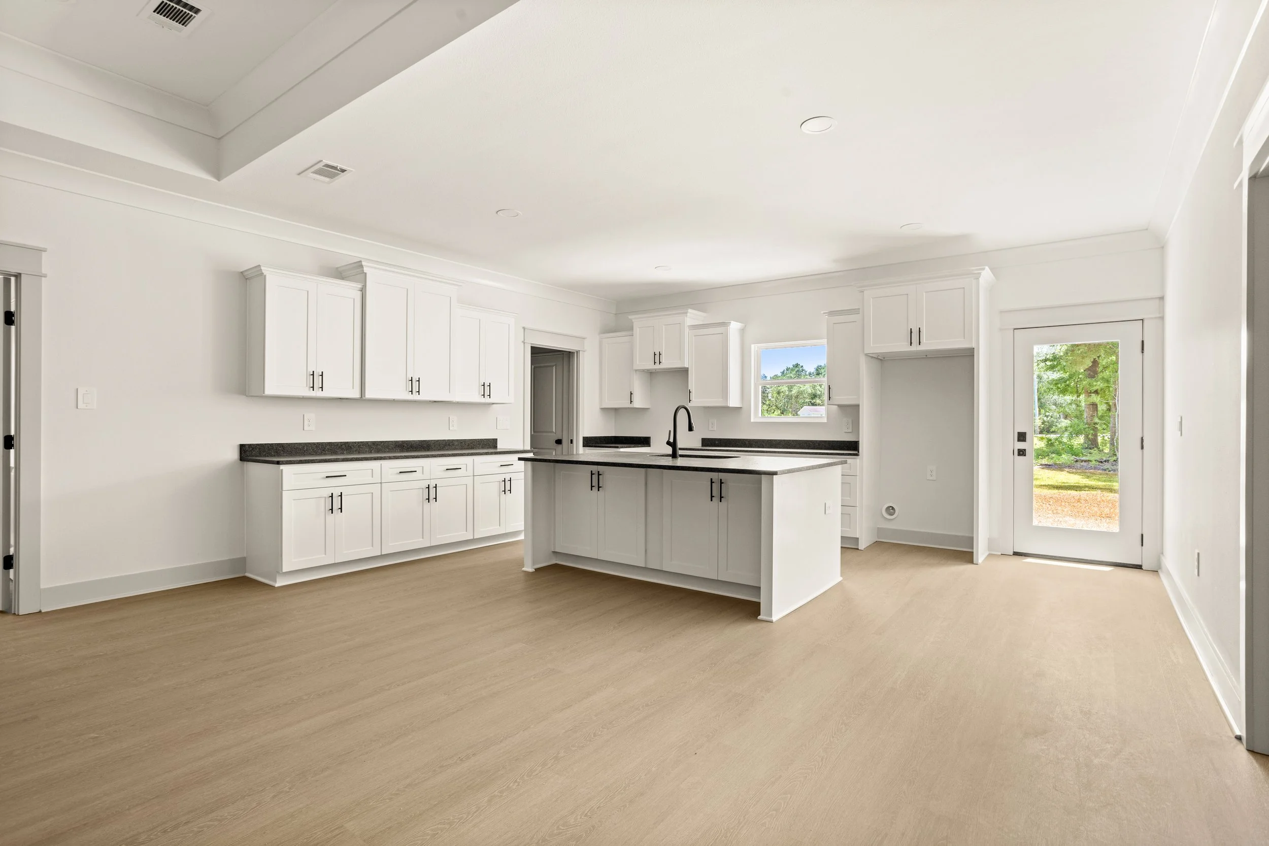 Empty, modern kitchen with white cabinets, black countertops, a kitchen island with a sink, and light wood flooring. There are windows and a door leading outside.