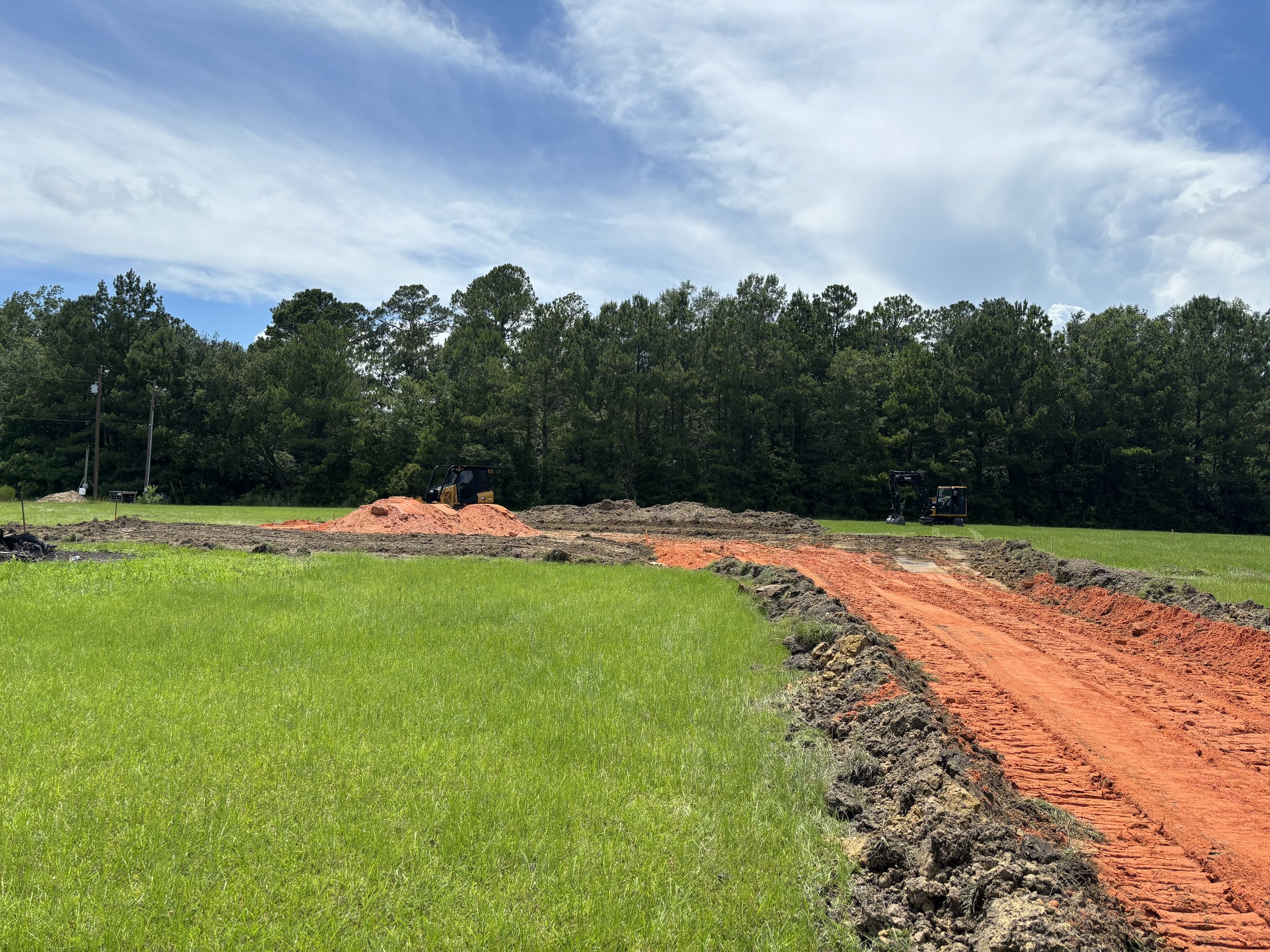 Construction site with a dirt road and small excavators working in a grassy field with trees in the background.