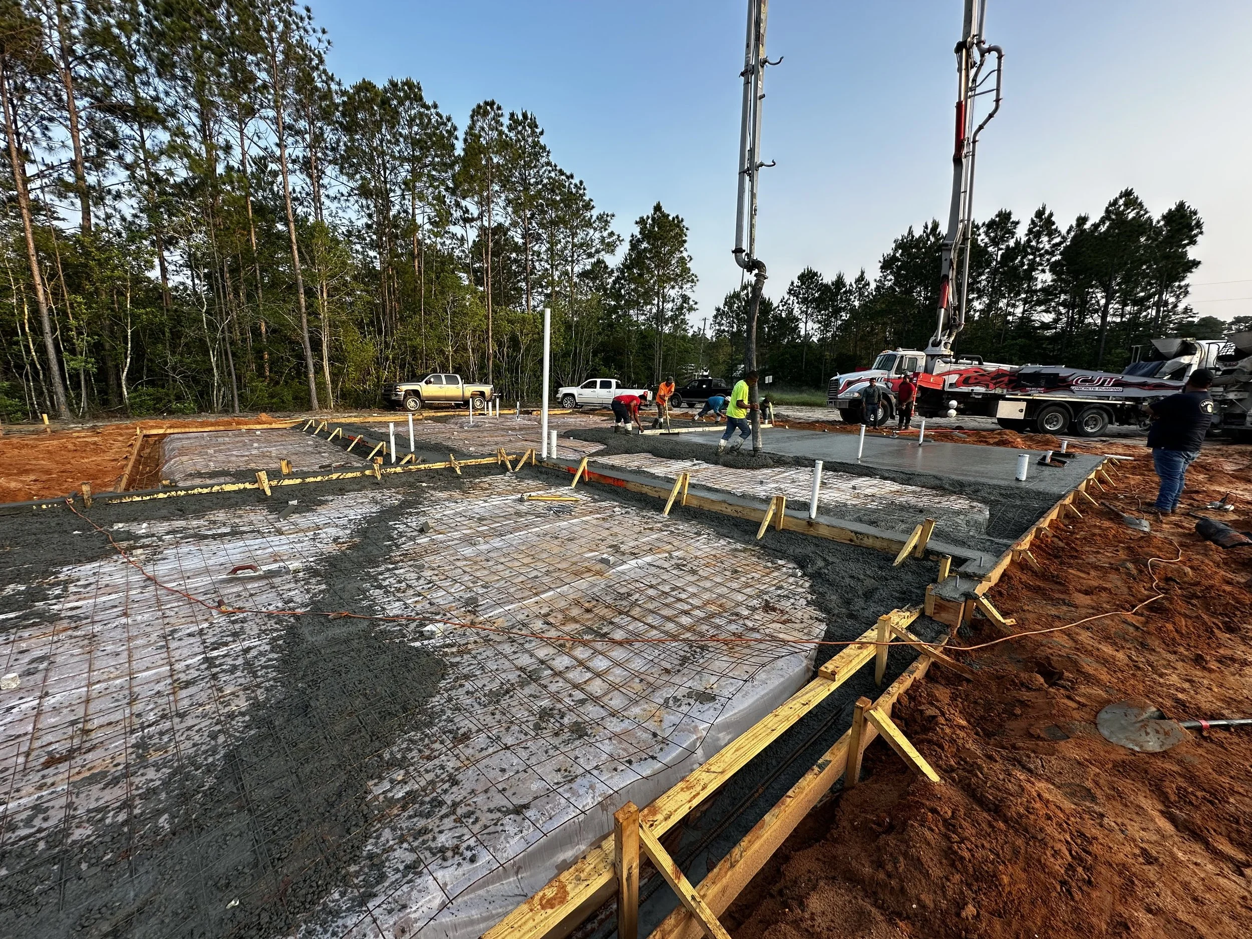 Construction workers pouring concrete for a building foundation with rebar, wooden formwork, and construction vehicles on a dirt site surrounded by trees.