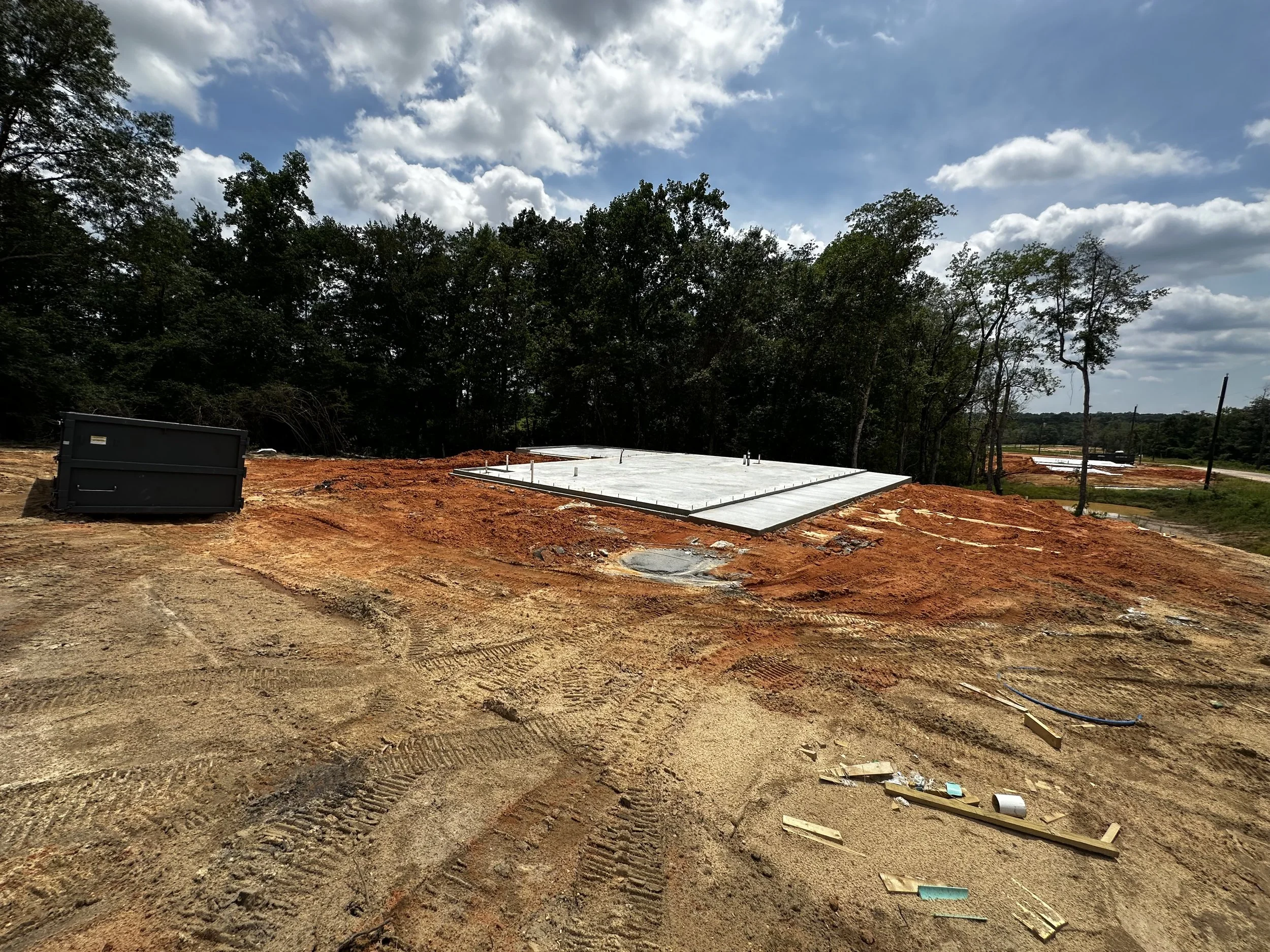 Construction site with a concrete foundation slab on reddish dirt, surrounded by trees and a partly cloudy sky.