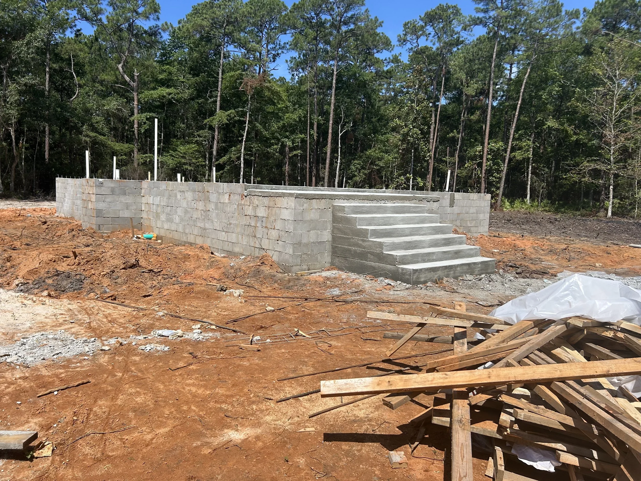 Construction site with a concrete block wall and stairs, surrounded by red dirt and construction debris, with a forest background.