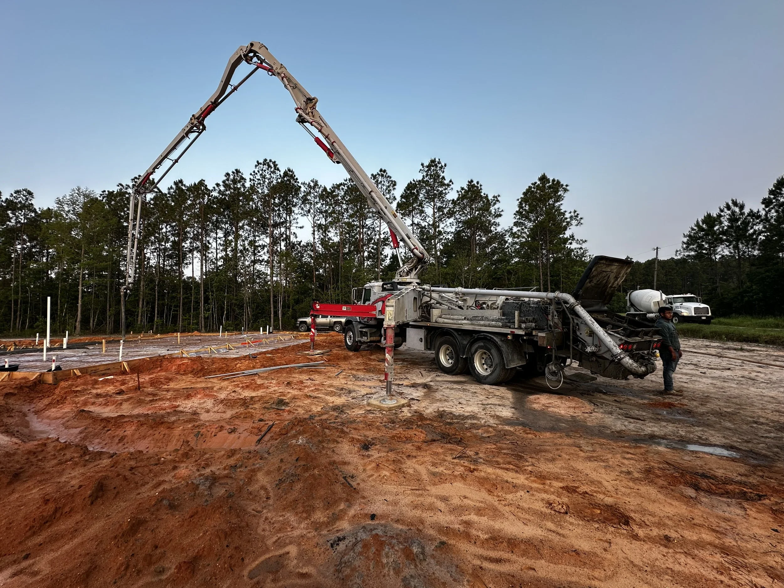 Construction site with a concrete pump truck pouring concrete onto a foundation, with a worker standing nearby and a forest in the background.