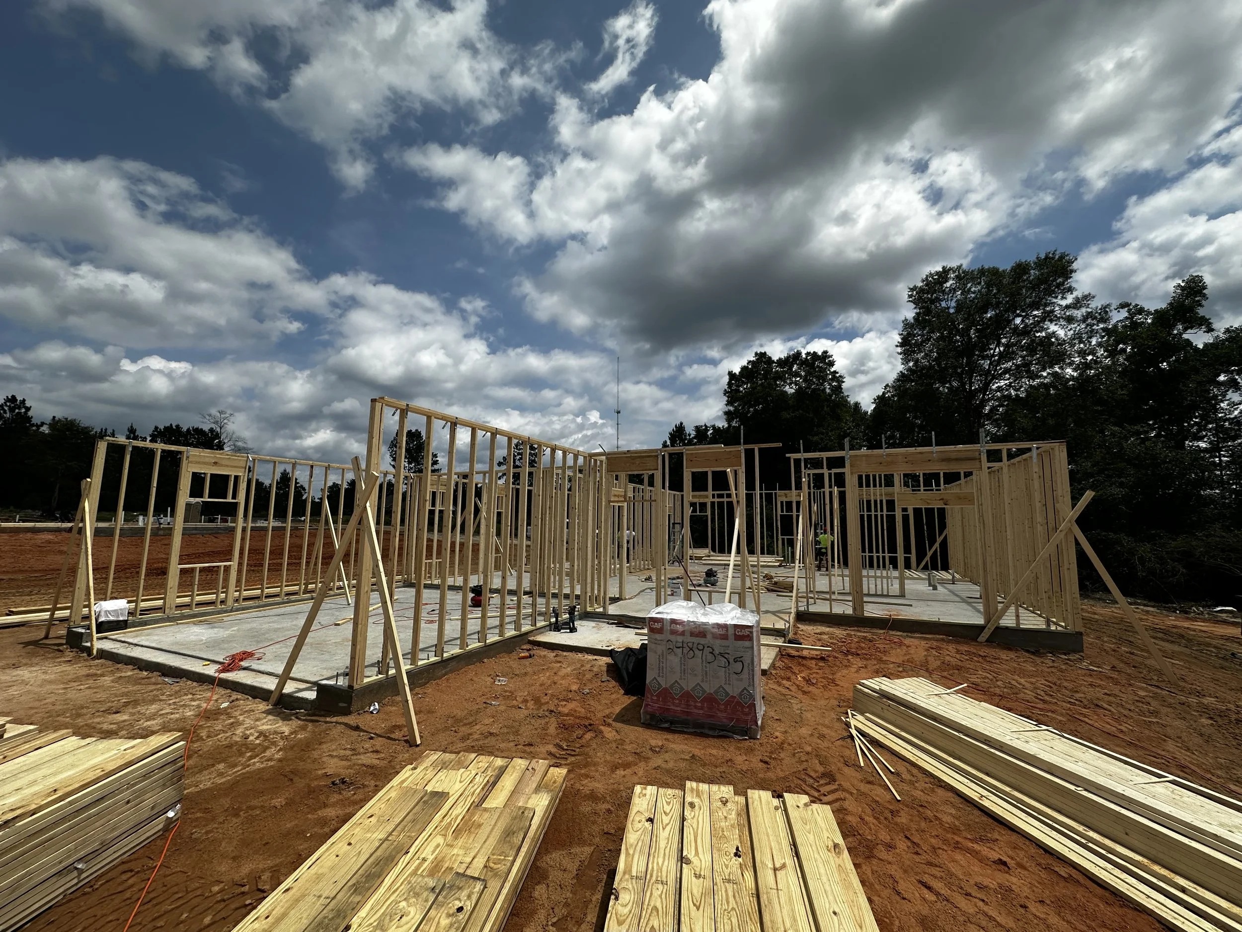Wood framing for a house under construction on a dirt site, with blue sky and scattered clouds overhead.