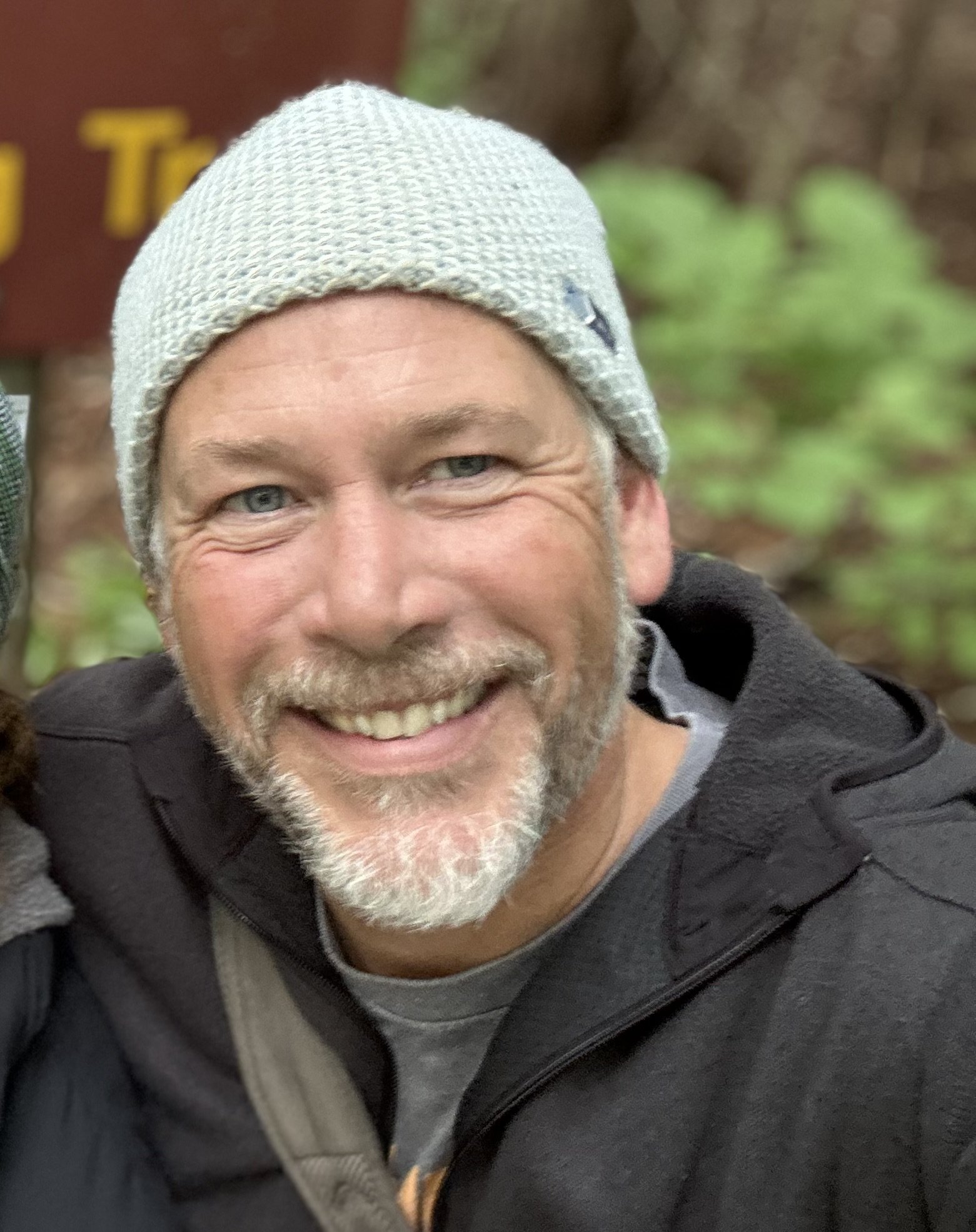 A smiling man with a gray beard and blue eyes wearing a light gray knit cap and black jacket outdoors.