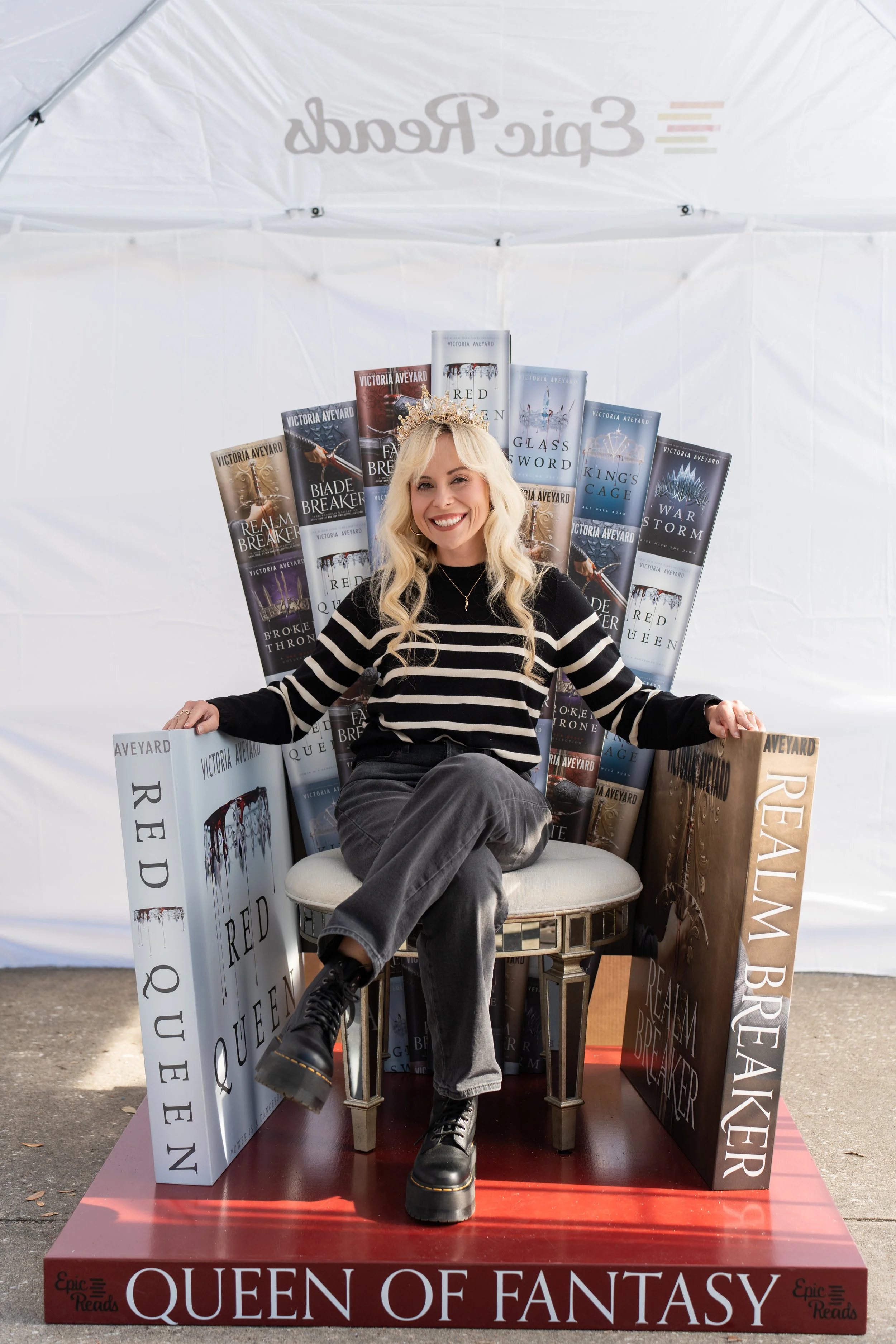 A fan perches on a throne made of oversized copies of books by Victoria Aveyard