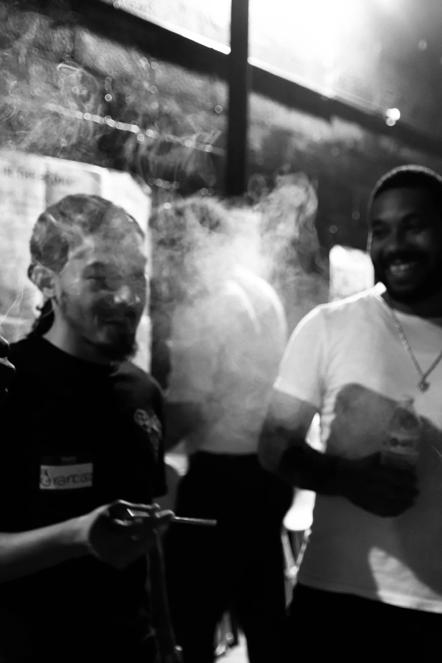 Two smiling men enjoying a moment together, one holding a cigarette, in a smoky indoor setting with a window in the background.