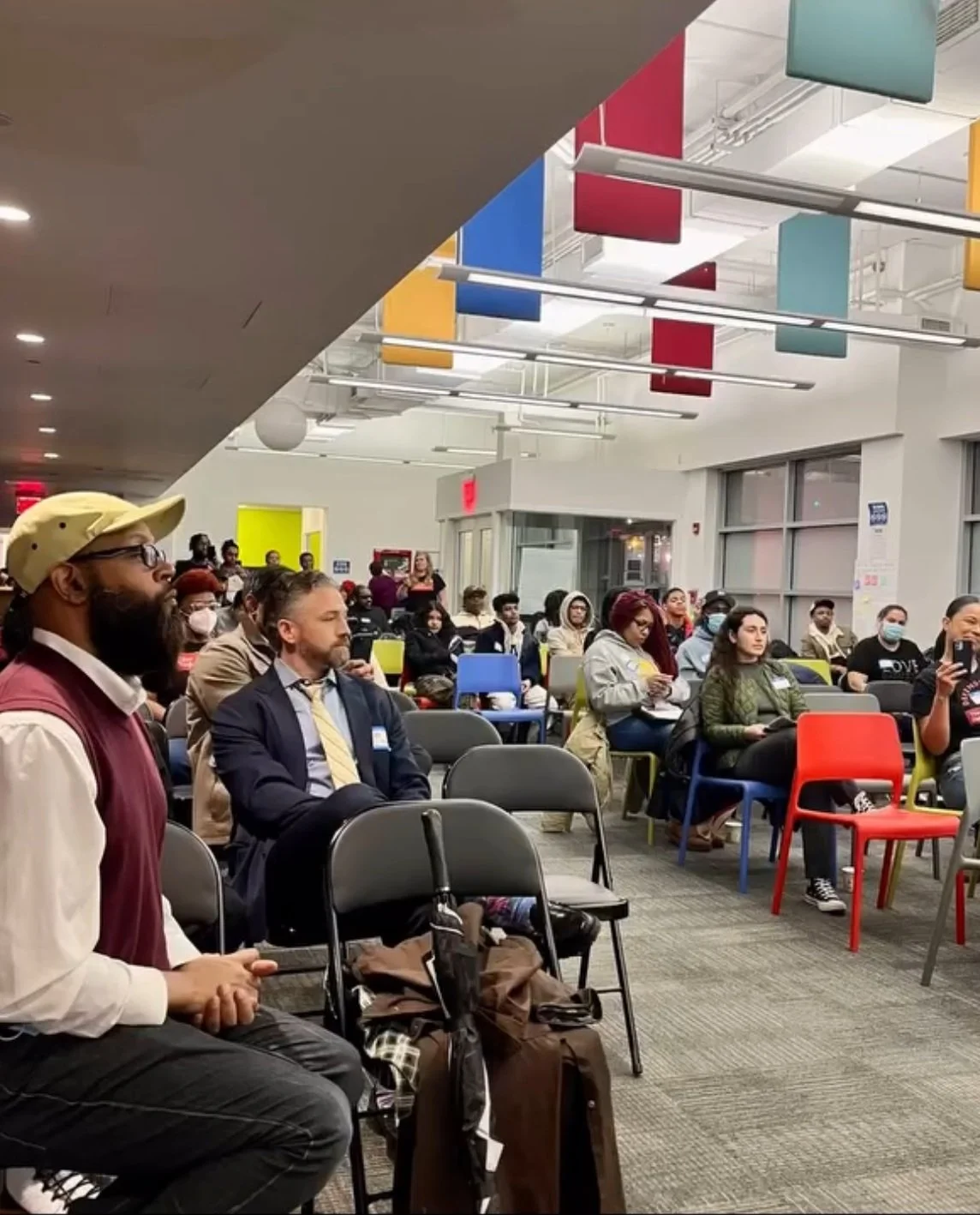 People attending a conference or event seated on chairs in a brightly lit room with colorful banners hanging from the ceiling.