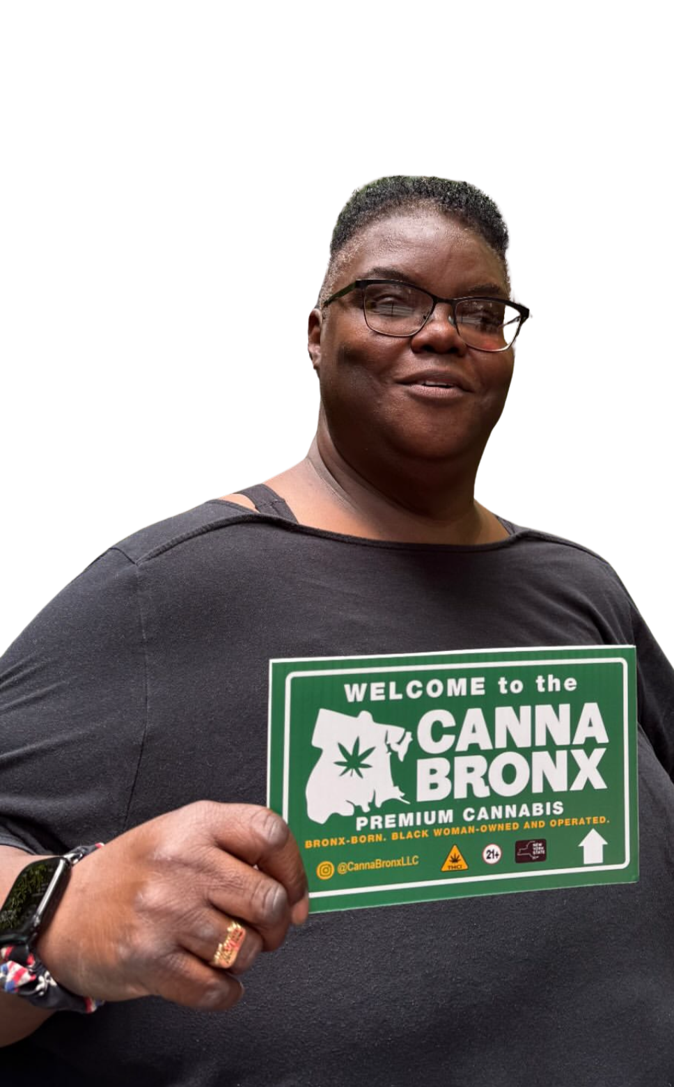 A person holding a sign that reads 'Welcome to the Canna Bronx, Premium Cannabis, Bronx-born, Black woman-owned and operated.' The individual is wearing glasses, a black t-shirt, and a bracelet.