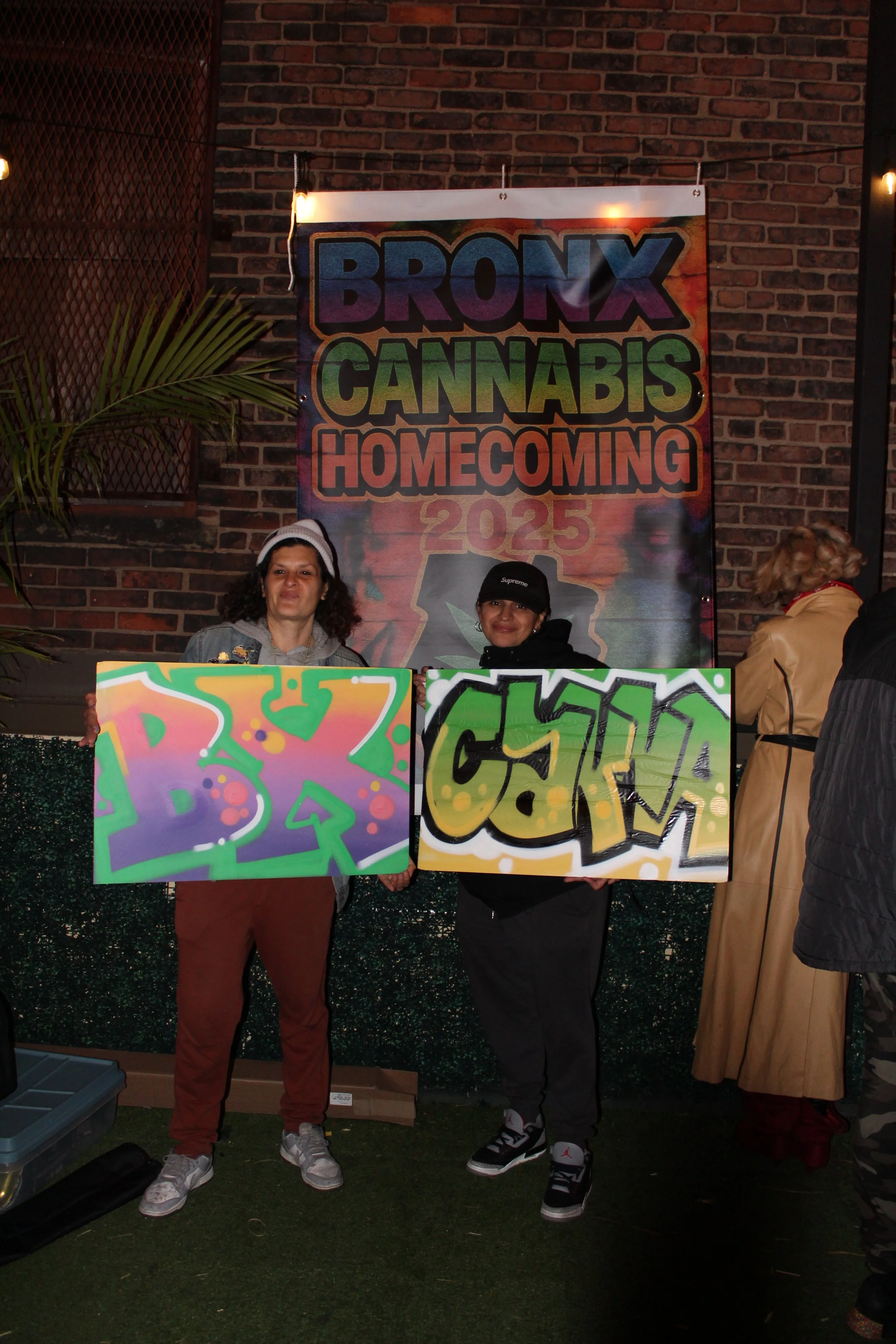 Two women holding spray-painted signs at the Bronx Cannabis Homecoming 2023 event, with a colorful banner in the background that says 'Bronx Cannabis Homecoming 2023' against a brick wall.