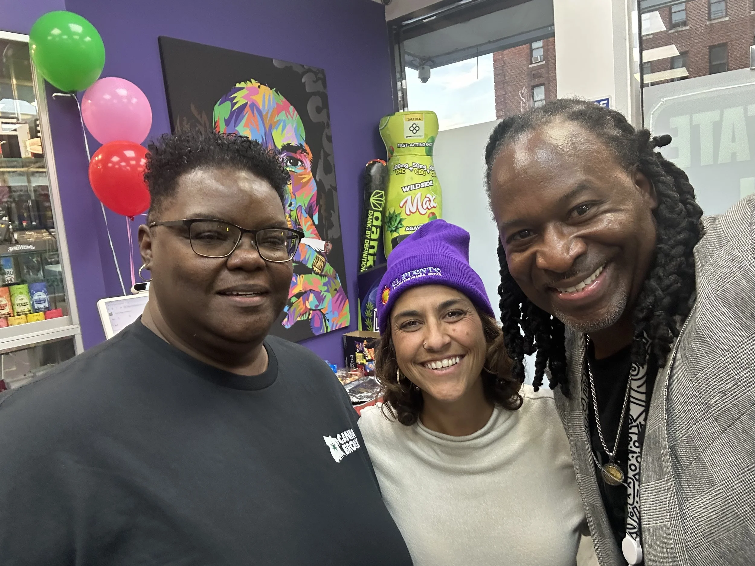 Three smiling people taking a selfie inside a colorful store with purple walls, balloons, and artwork in the background.