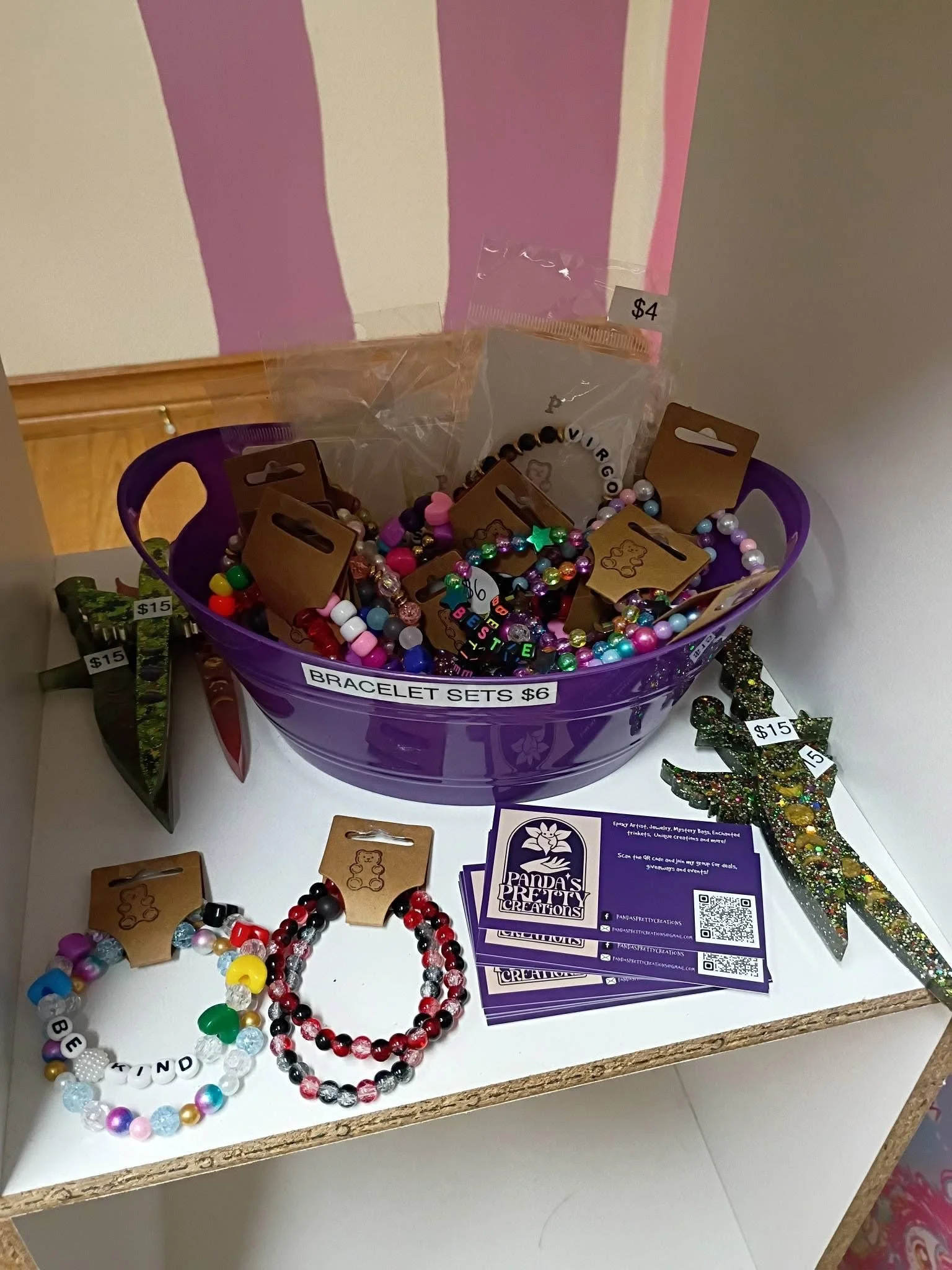 bucket of handmade beaded bracelets , resin glitter daggers displayed on white shelf next to bucket