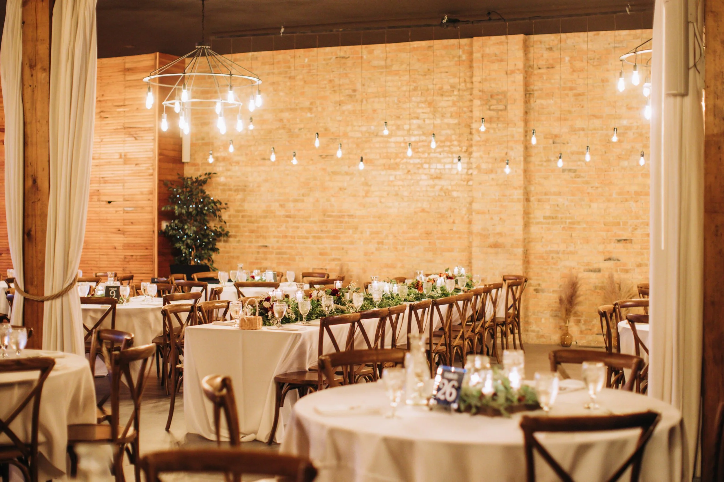 Wedding reception area with round and rectangular tables adorned with white tablecloths, floral centerpieces, candles, and glassware, set against a backdrop of a brick wall and string lights hanging from the ceiling.