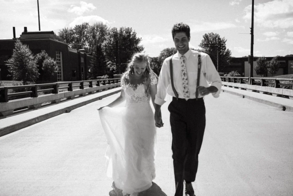 A black and white photo of a smiling couple holding hands while walking on a bridge, with trees and buildings in the background.
