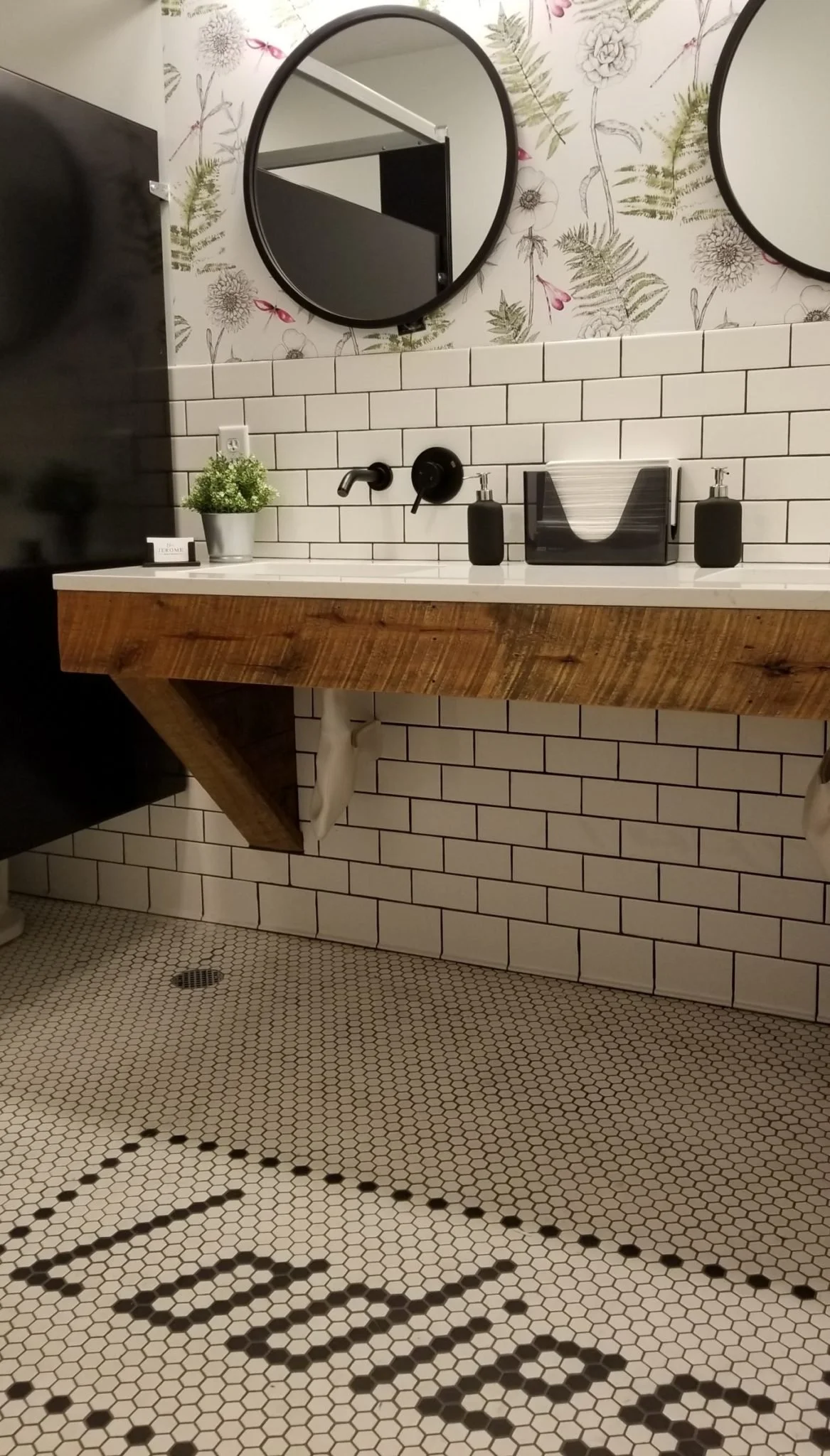 Modern bathroom with white subway tile walls, a wooden floating vanity, two round mirrors, a black faucet, soap dispensers, a tissue box, a potted plant, and a black and white hexagon tiled floor with the word 'HELLO' spelled out with black tiles.