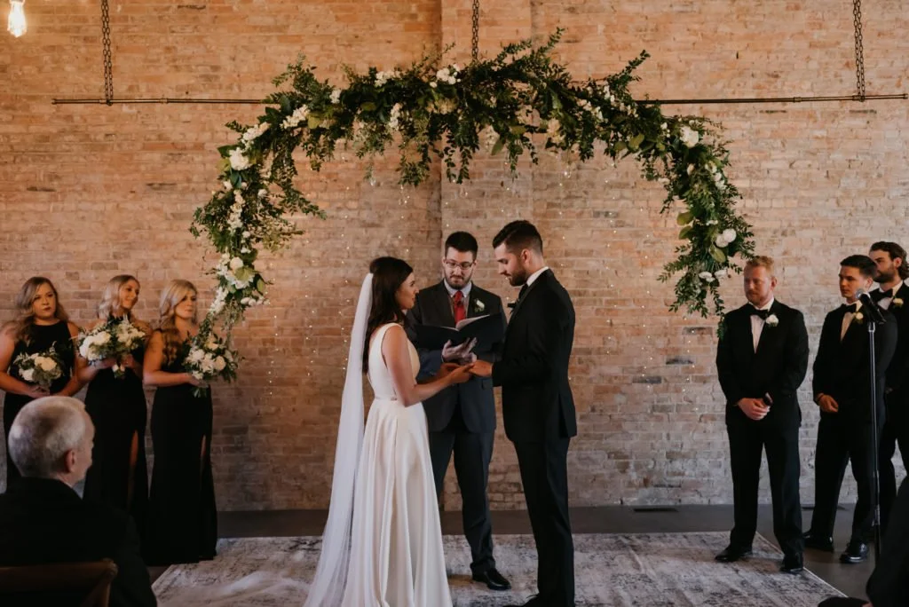 Bride and groom having a wedding ceremony under a floral arch in front of a brick wall, with bridesmaids in black dresses holding bouquets on the left and groomsmen in black suits on the right.