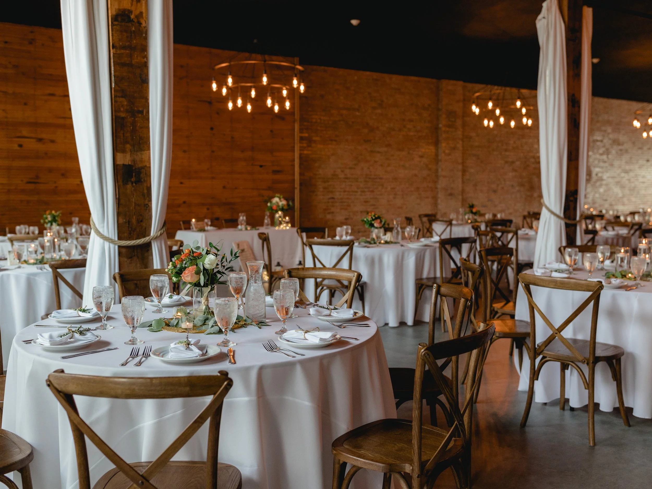 Elegant event space set with round tables covered in white tablecloths, decorated with floral centerpieces, candles, glassware, and dinnerware, with wooden chairs, brick walls, and warm lighting.
