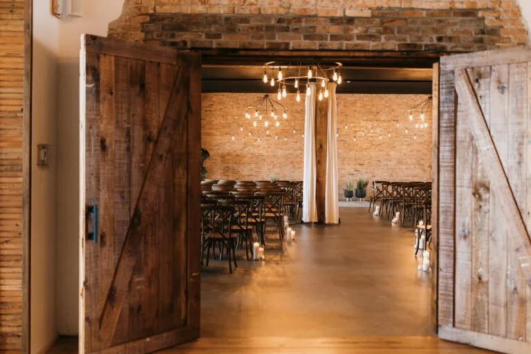 Wedding or event reception area with rustic decor, wooden tables and chairs, exposed brick walls, hanging string lights, and candles along the floor.