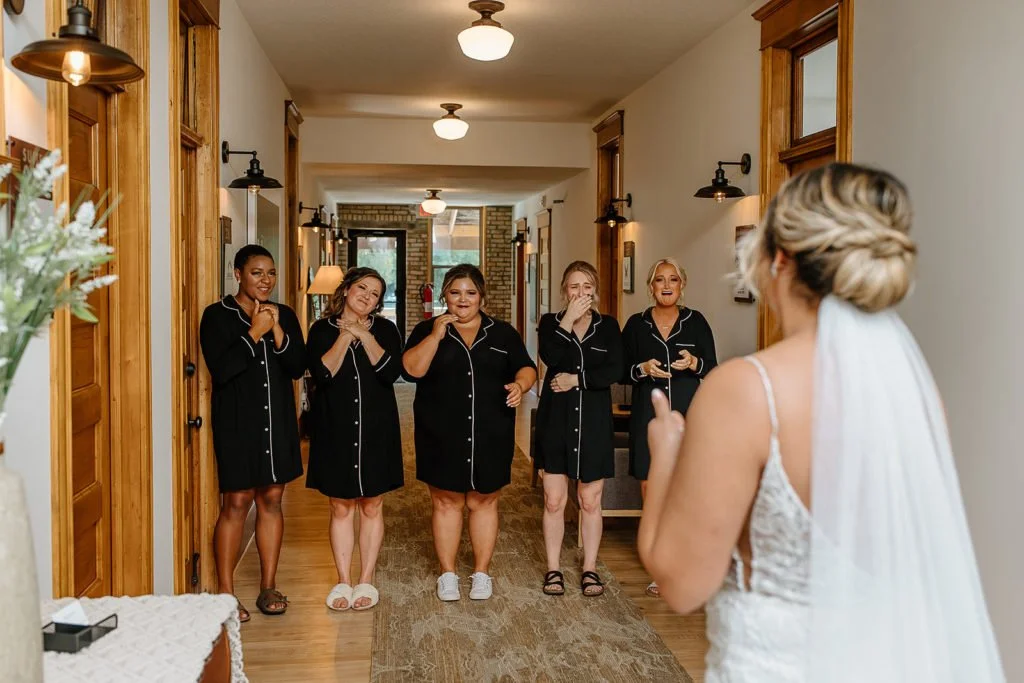 Bride in white wedding dress with veil holding a phone, looking at five women with black pajamas, reacting emotionally; inside a warmly lit hallway in a house.