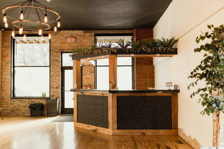 Empty reception or bar area with brick wall, large windows, hanging Edison-style light bulbs, and a wooden counter with greenery on top.