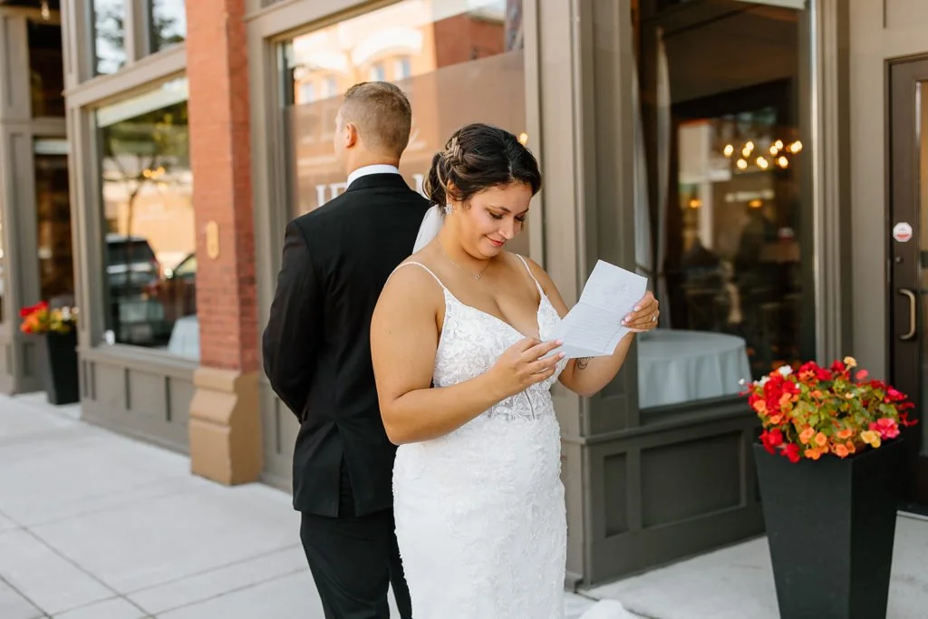 A bride in a white wedding dress and a groom in a black suit are standing outside a building, with the bride reading a note or letter.
