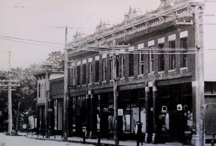 Black and white photo of an old two-story brick building with decorative architectural details, storefronts on the ground level, and power lines overhead.