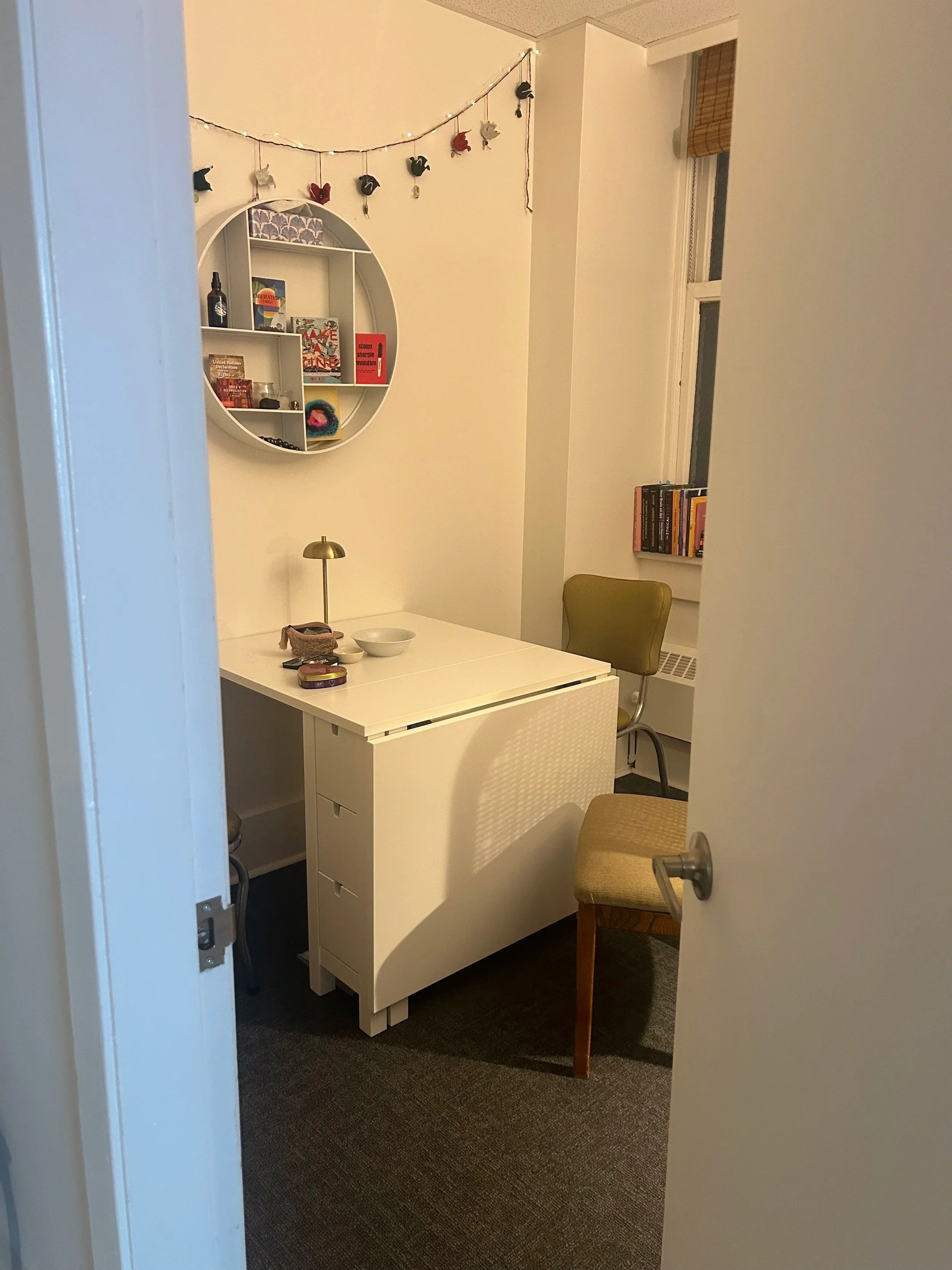 View of a small room with a white desk, beige chairs, a bookshelf, and decorative wall hanging, seen through a partially open door.
