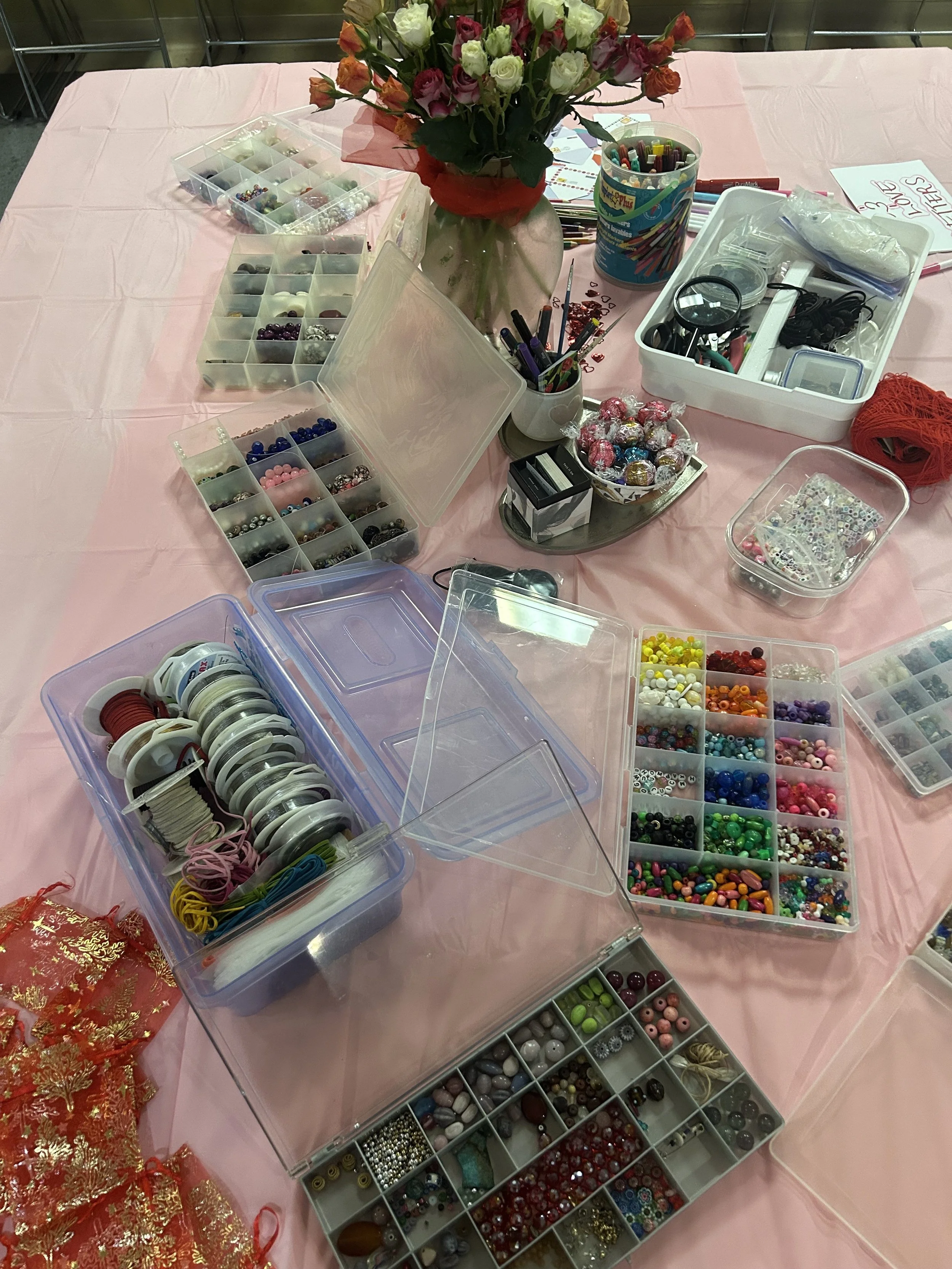 A table covered with a pink tablecloth displays various containers filled with colorful beads, buttons, ribbons, and craft supplies. In the center, there is a vase of multicolored flowers.