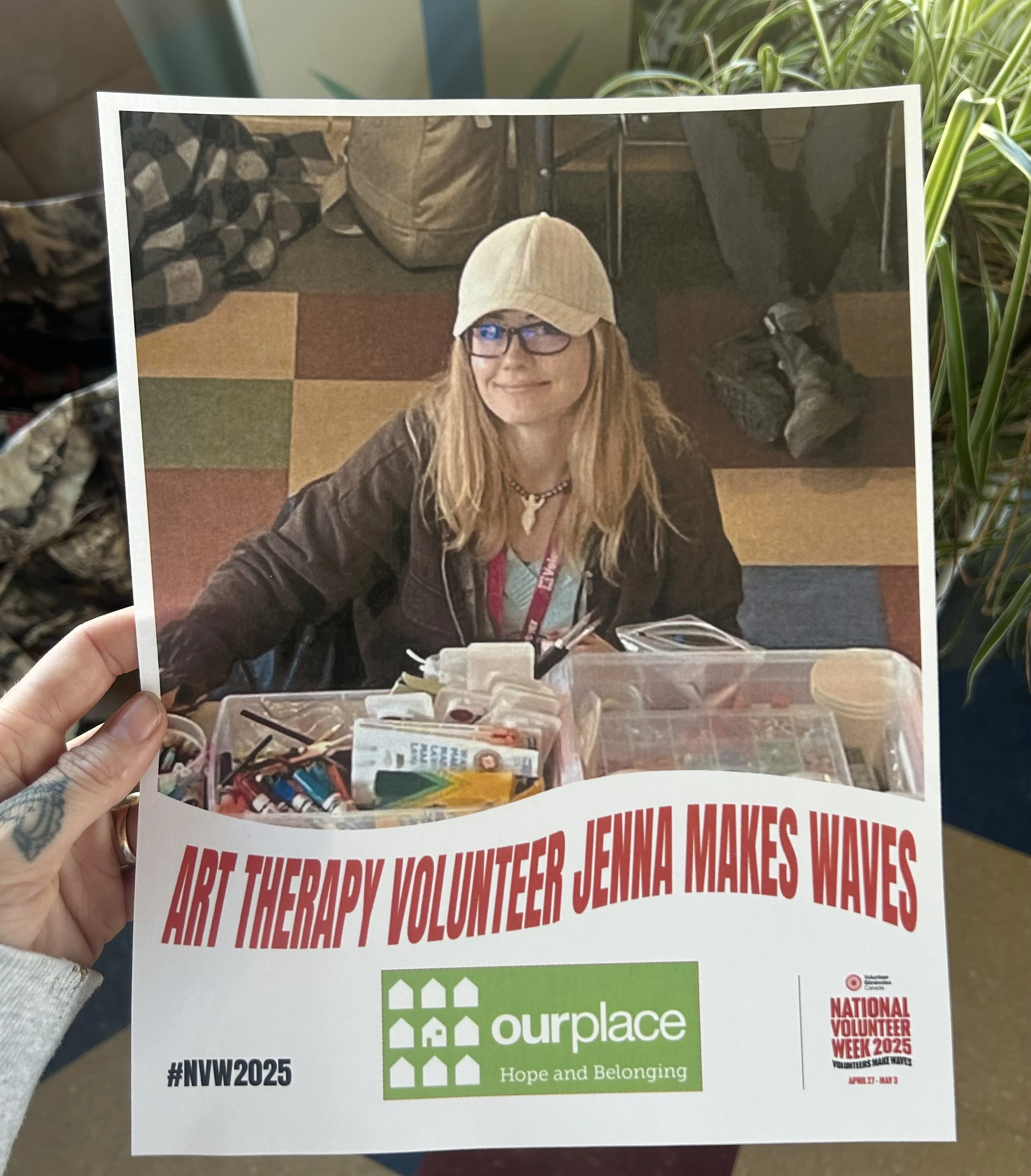 Photograph of a printed flyer held in hand, showing an image of a woman wearing glasses and a beige cap, smiling, sitting at a table with various items, and with a background of colorful carpet tiles and shoes. The flyer promotes a volunteer event for the National Volunteer Week 2025, with the hashtag #NVW2025 and logo for 'ourplace' community organization, and the slogan 'Hope and Belonging'.