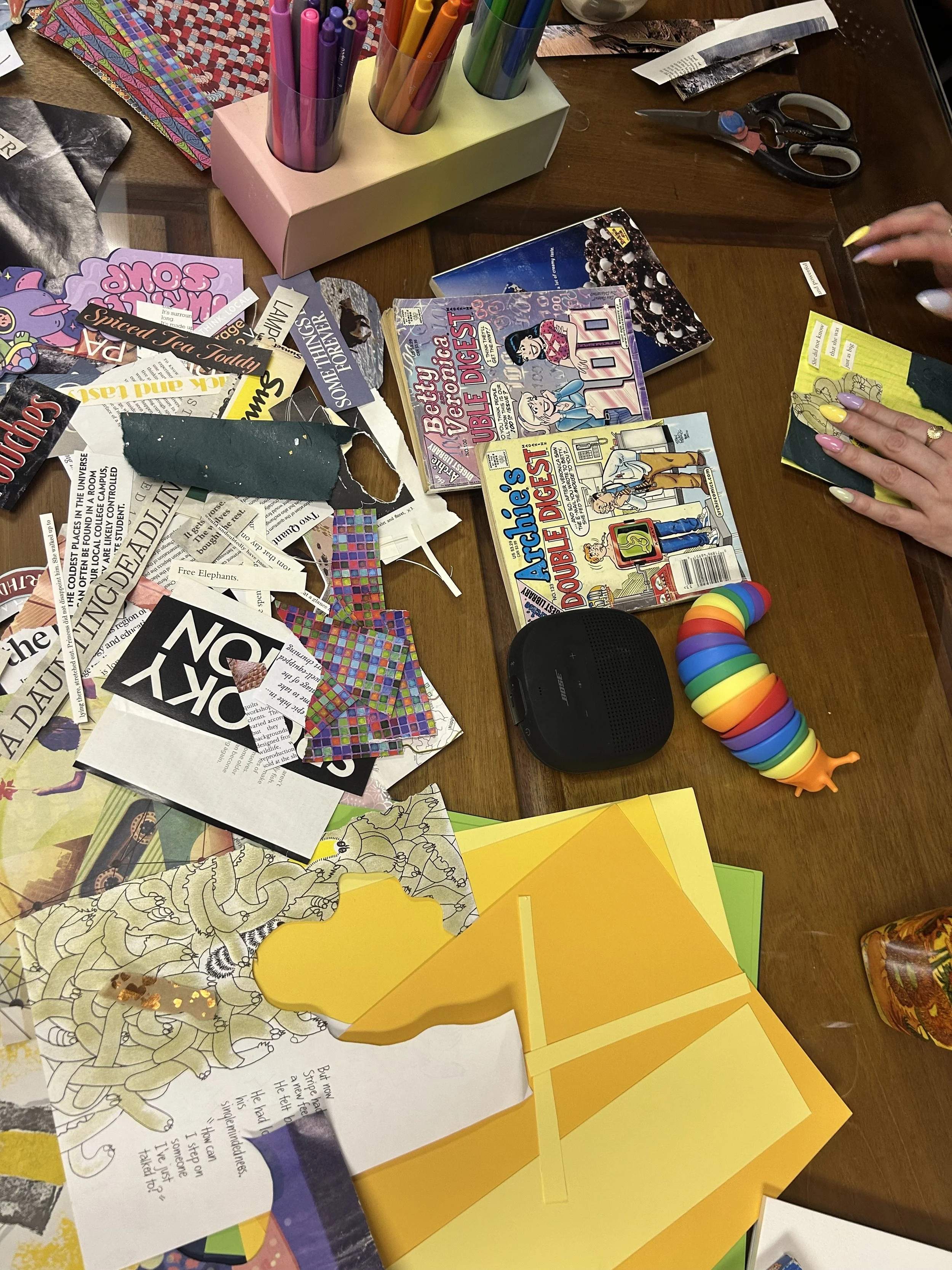 Table scattered with paper crafts, coloring pages, and various stationery items including colored paper, sticky notes, and scissors. There is a colorful rainbow spiral toy, a Bose speaker, and a container of colored markers arranged in holders. A person's hands are visible, involved in a craft activity.