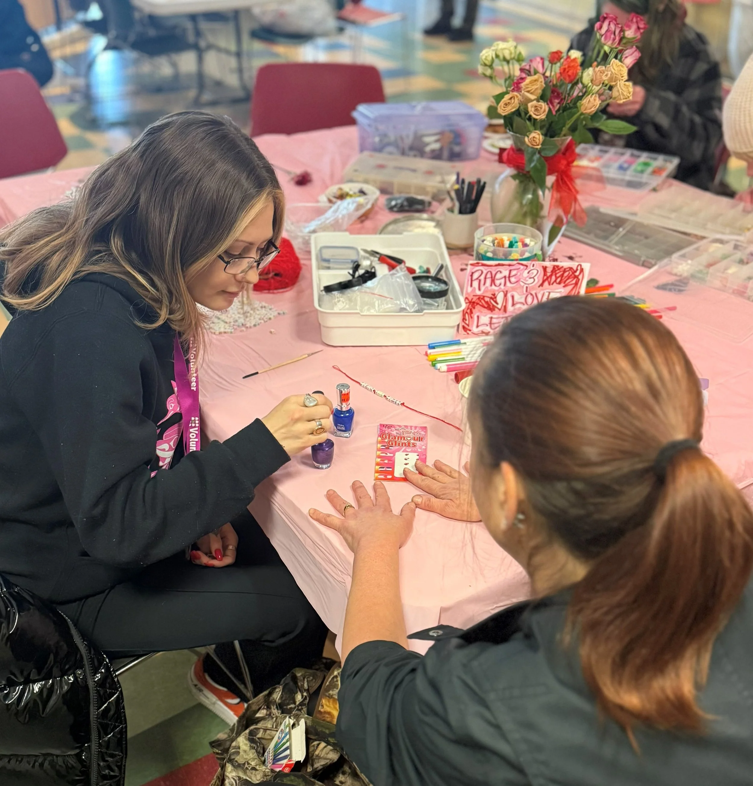 People sitting around a table engaged in a craft activity, with a large vase of multicolored roses as the centerpiece and various crafting supplies scattered on the pink tablecloth.