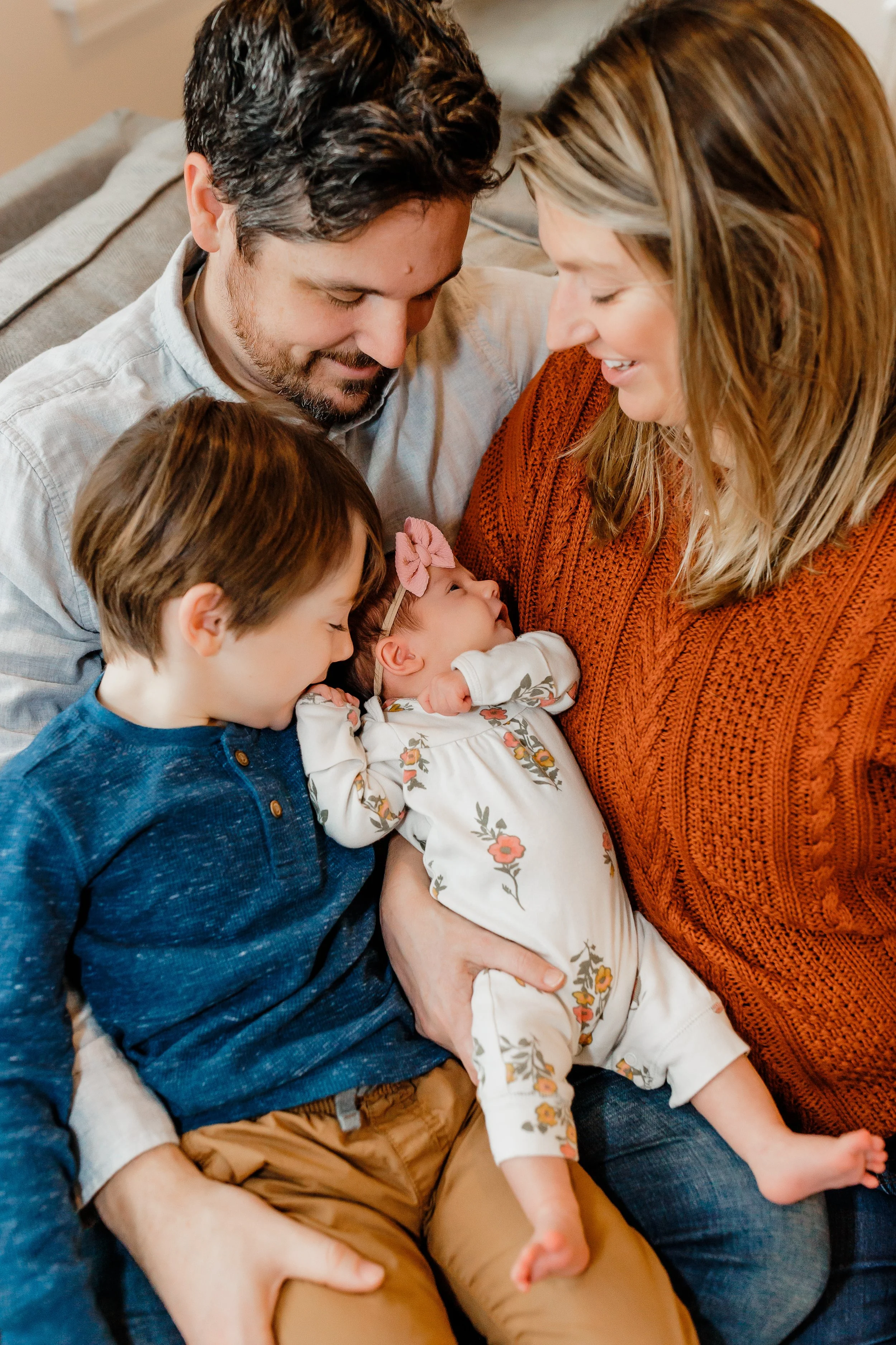 A family of four, including a man, a woman, a young boy, and a newborn baby, gathered closely together on a couch, smiling and looking lovingly at the baby.