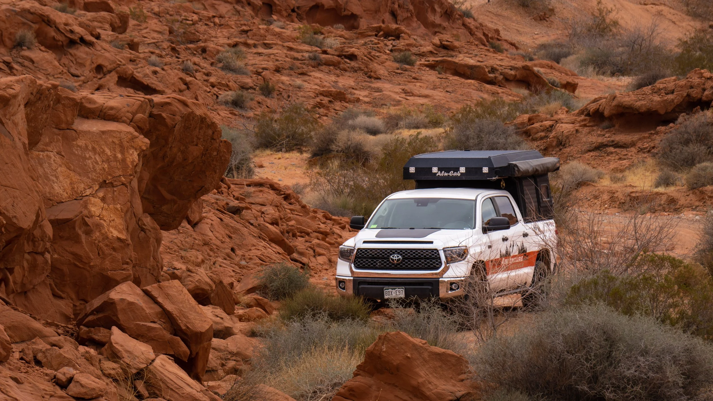 Capture those epic 4x4 overland trips.  A white Toyota vehicle with a rooftop camper is parked on rough, red rocky terrain in a desert landscape, at valley of fire state park.